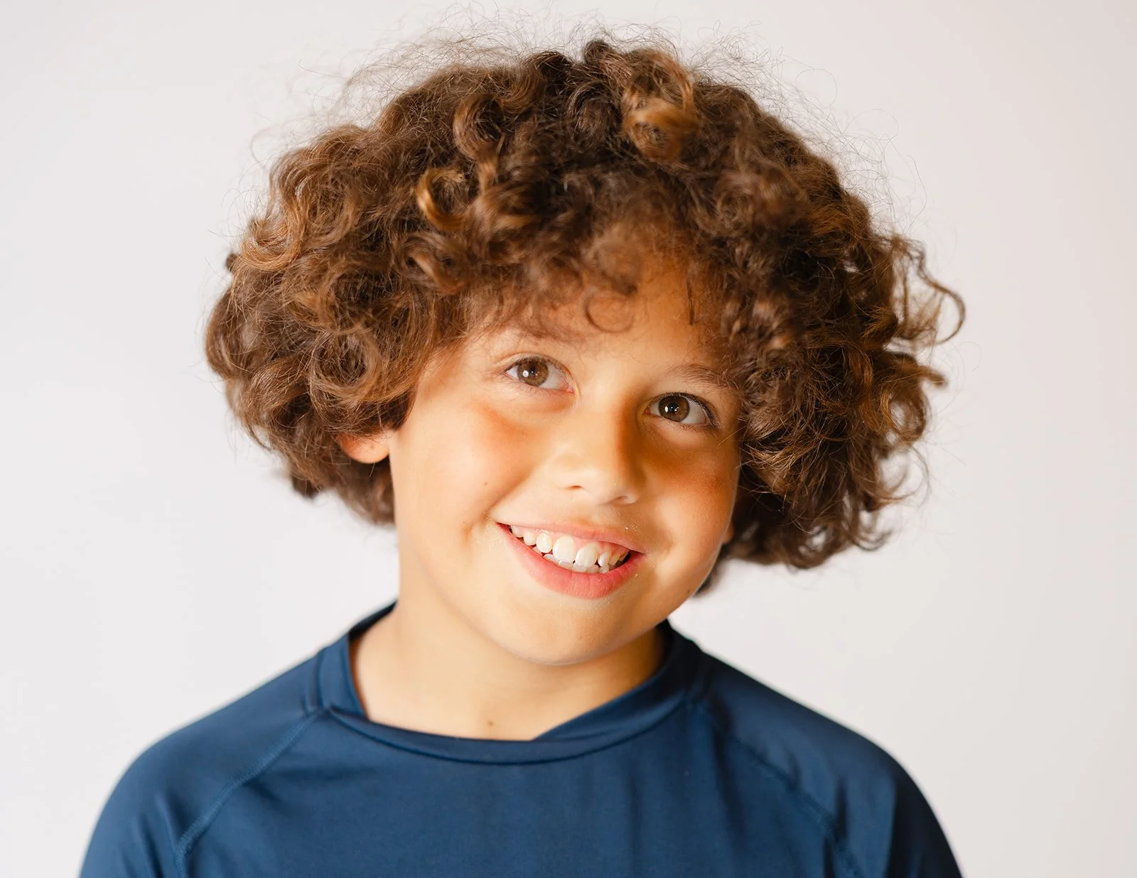 Close-up of a boy with curly brown hair, wearing a navy blue shirt, smiling against a plain white background.