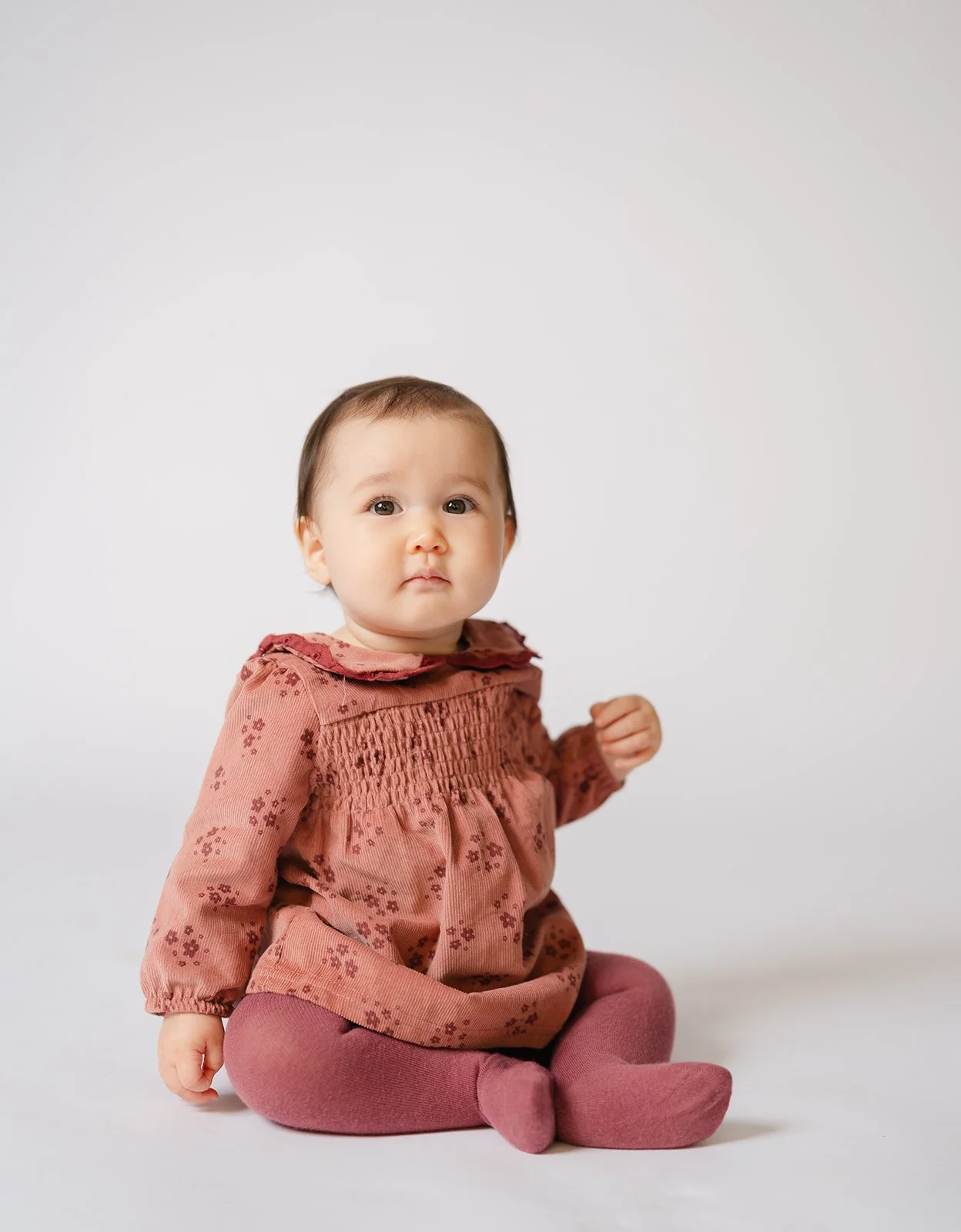 A baby girl sitting on a white background, wearing a rust-colored dress with floral patterns and pink tights.