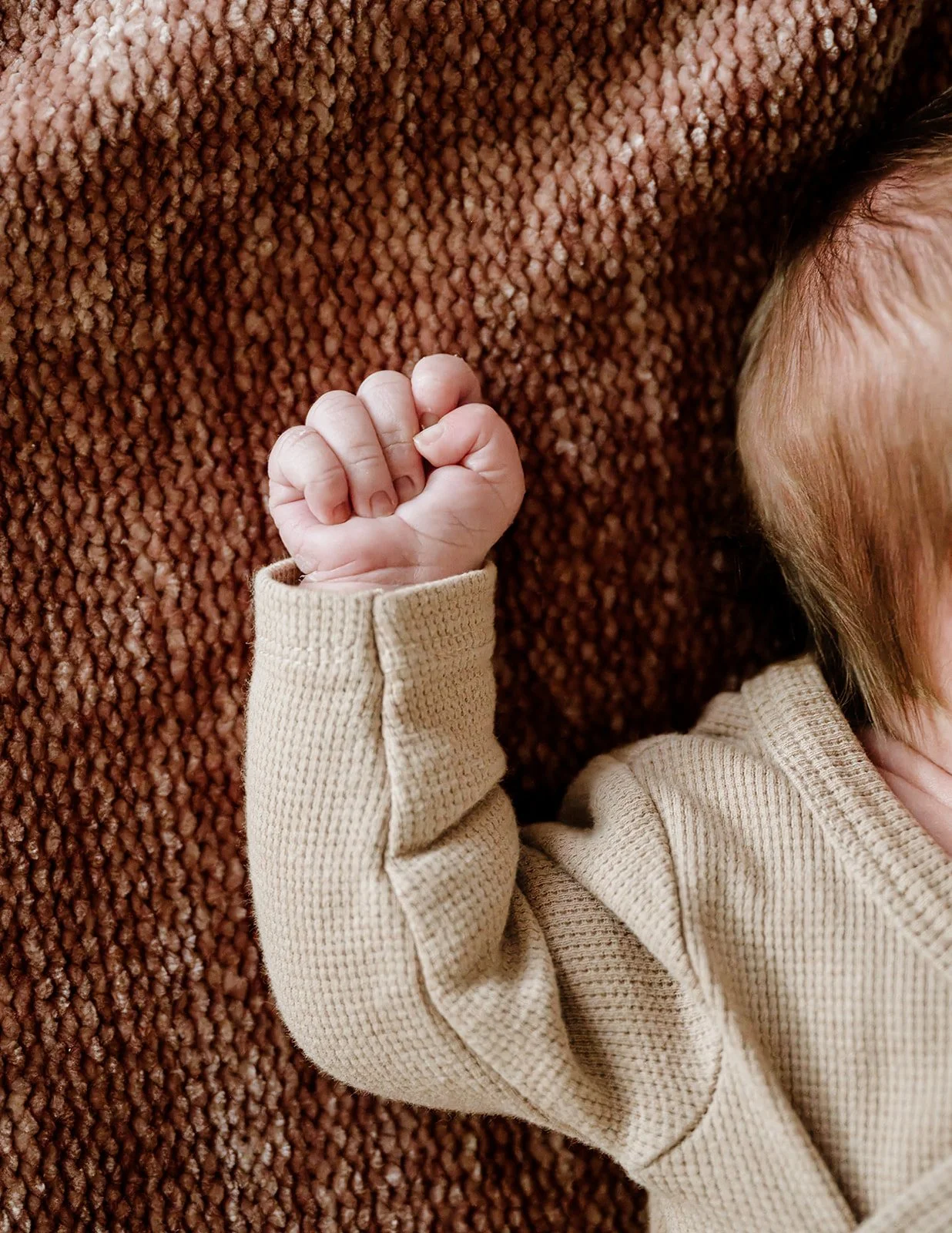 Close-up of a baby's hand clenched into a fist, with a cozy beige sweater sleeve showing, resting on a textured brown blanket. Part of a child's face with light-colored hair is visible on the right.