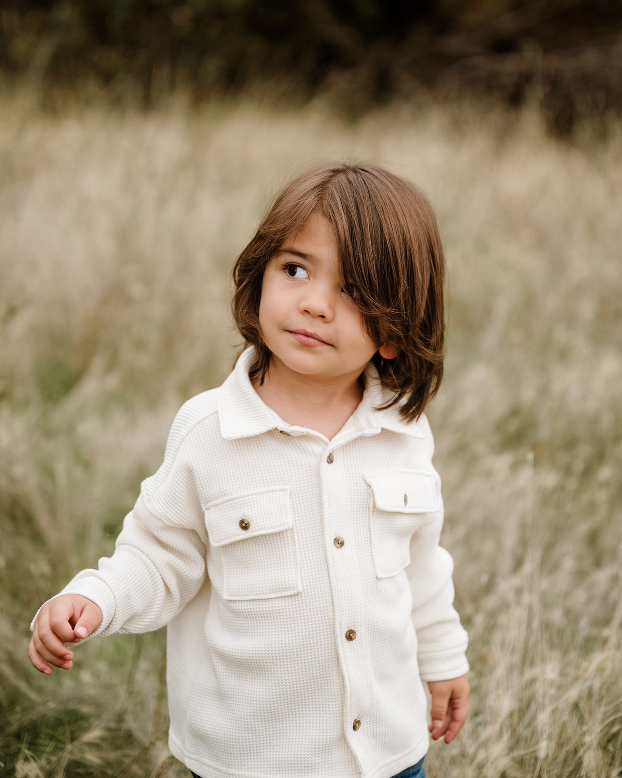 A young child with shoulder-length brown hair wearing a cream-colored shirt standing outdoors in a grassy field.