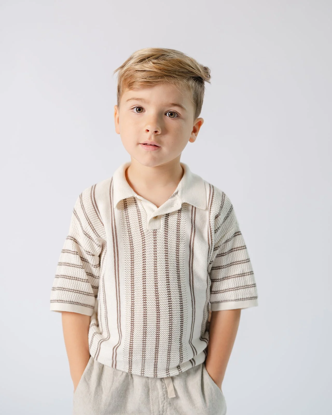 A young boy with light brown hair and light skin, wearing a cream-colored polo shirt with brown stripes, standing against a plain white background.