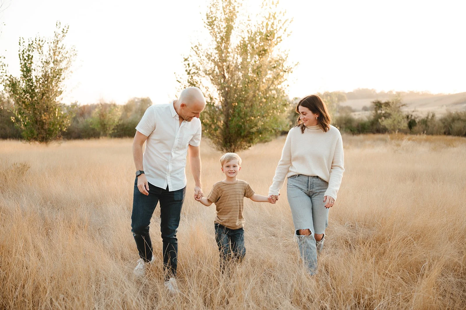 A family of three walking through a field of tall, golden grass during sunset, holding hands and smiling.