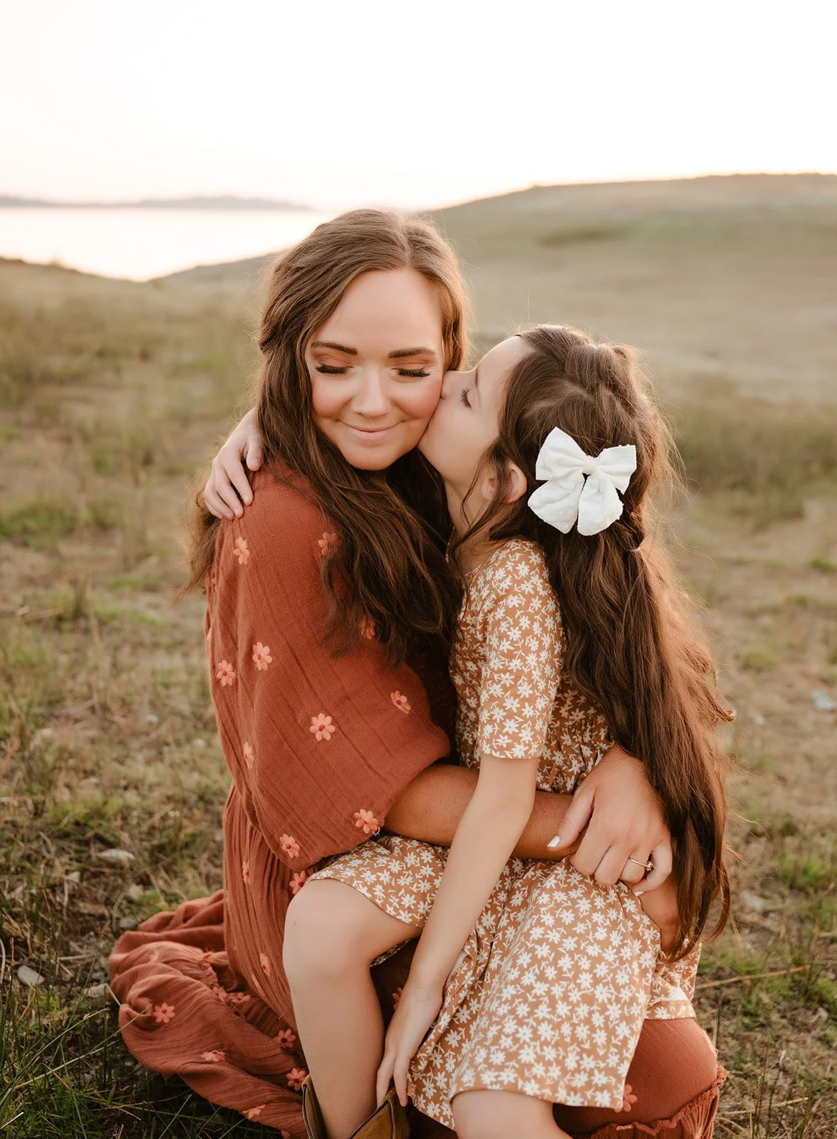 A woman and a young girl sitting outdoors on a grassy field during sunset, with the girl kissing the woman on the cheek.