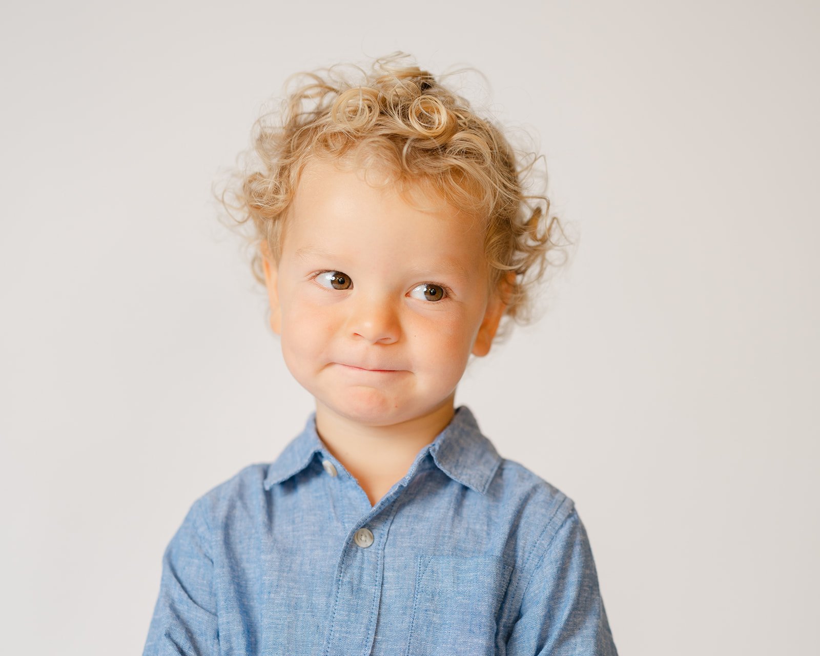 A young boy with curly blonde hair and light skin wearing a blue button-up shirt, looking to the side with a slight smile against a plain white background.