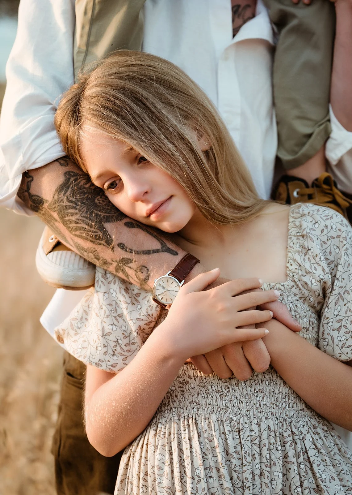 A young girl with blonde hair, wearing a floral dress, cuddles in the arms of an adult, whose arm with tattoos is wrapped around her, in an outdoor setting.