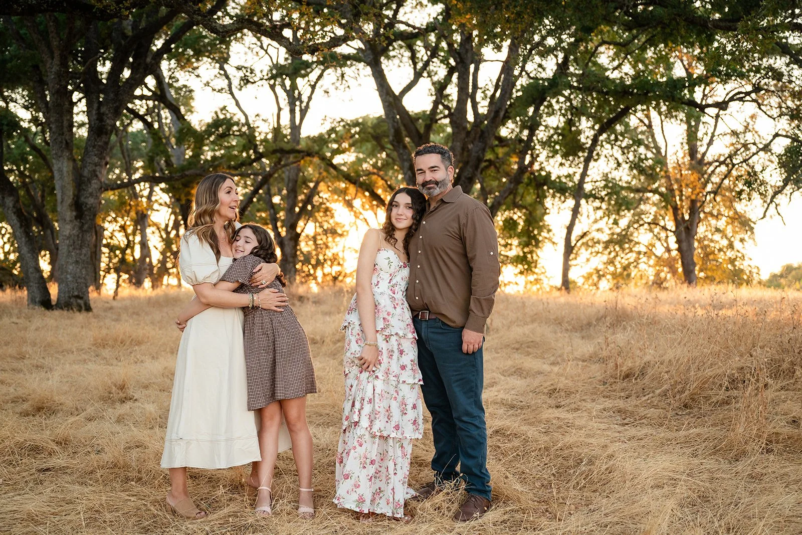 Family of four standing in a field of dry grass with tall trees in the background during sunset. The mother, with long wavy hair, is hugging a young girl in a checkered dress. The father, with a beard, is standing next to an older girl in a floral dr