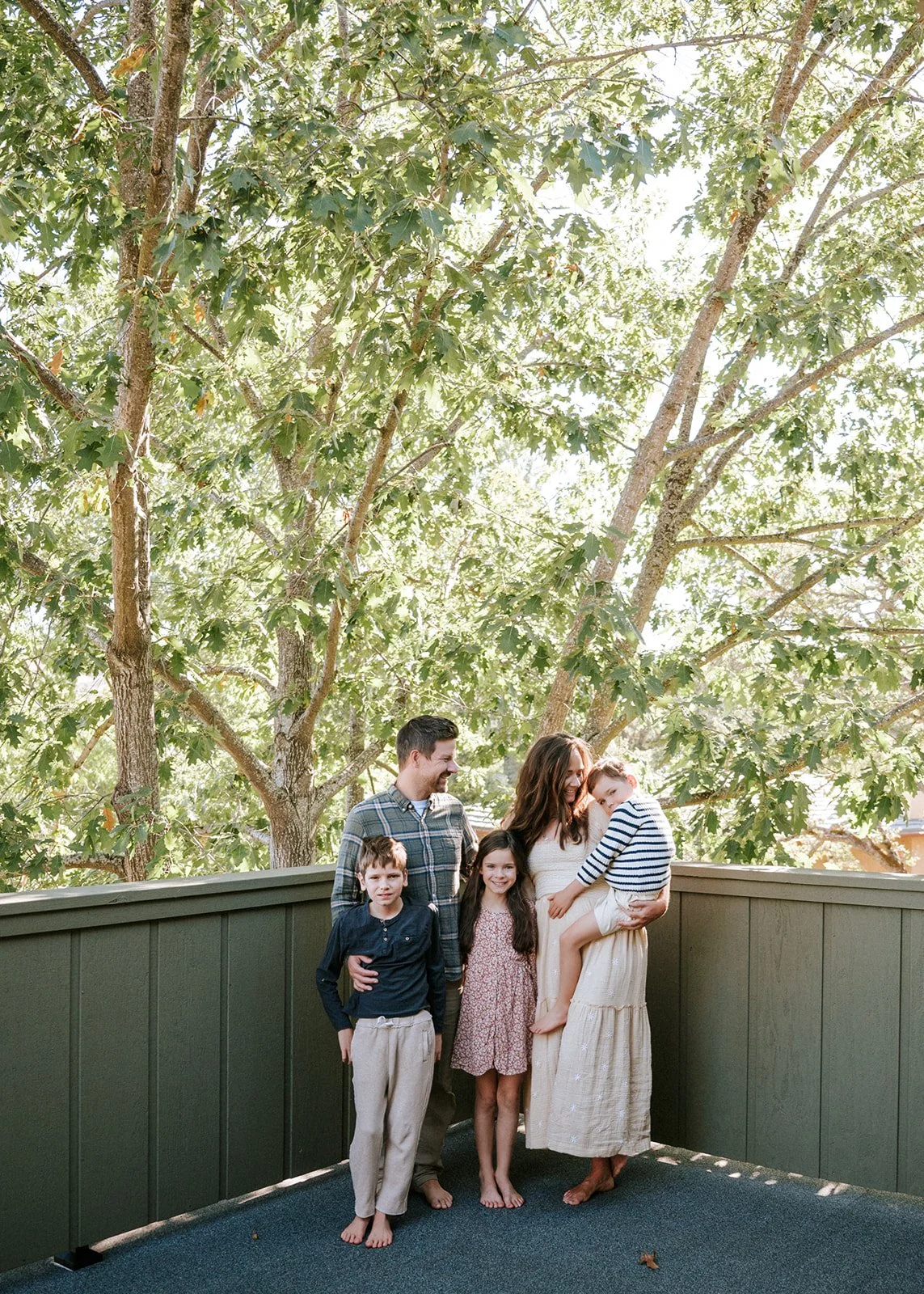 A family of five standing on a deck with a large tree behind them, smiling and interacting.