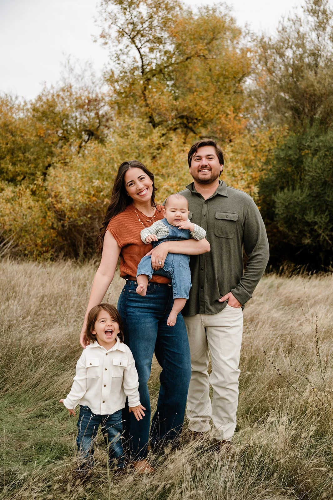 A family of four standing in a field of tall grass with autumn trees in the background. The mother has long dark hair, is smiling, and holding a baby boy dressed in denim overalls. The father has a beard, is smiling with hands in pockets, and is dres