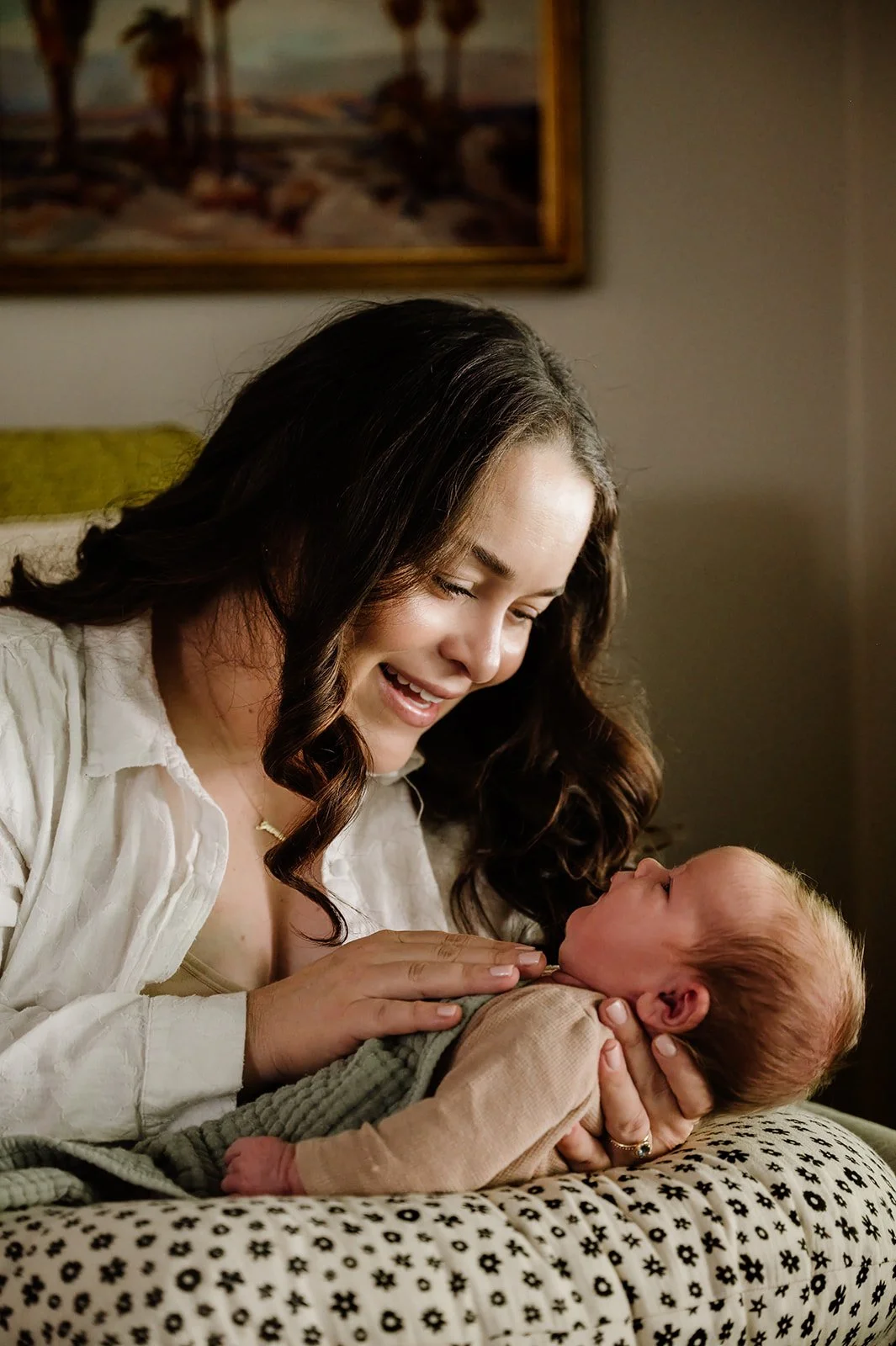 A woman with dark, wavy hair looking at a newborn baby lying on a patterned pillow, smiling warmly.