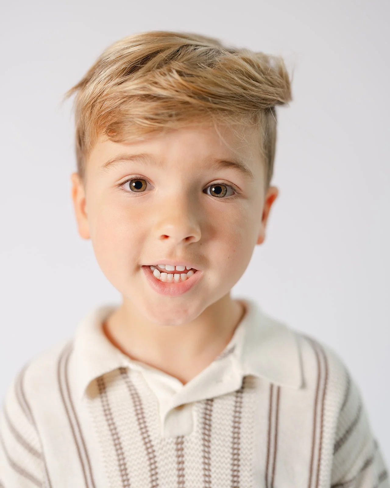 A young boy with light brown hair, hazel eyes, and a slight smile showing his teeth, wearing a beige and brown striped sweater, against a plain white background.