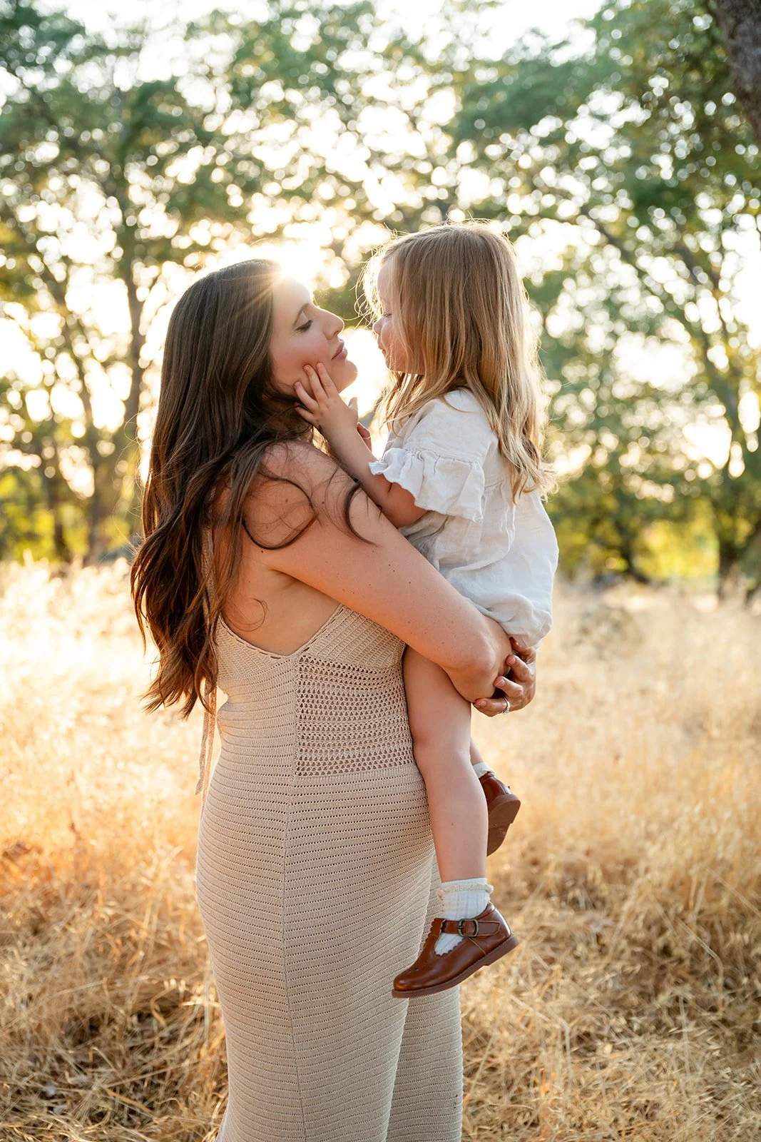 A woman holding a young girl in a field with trees in the background during sunset.
