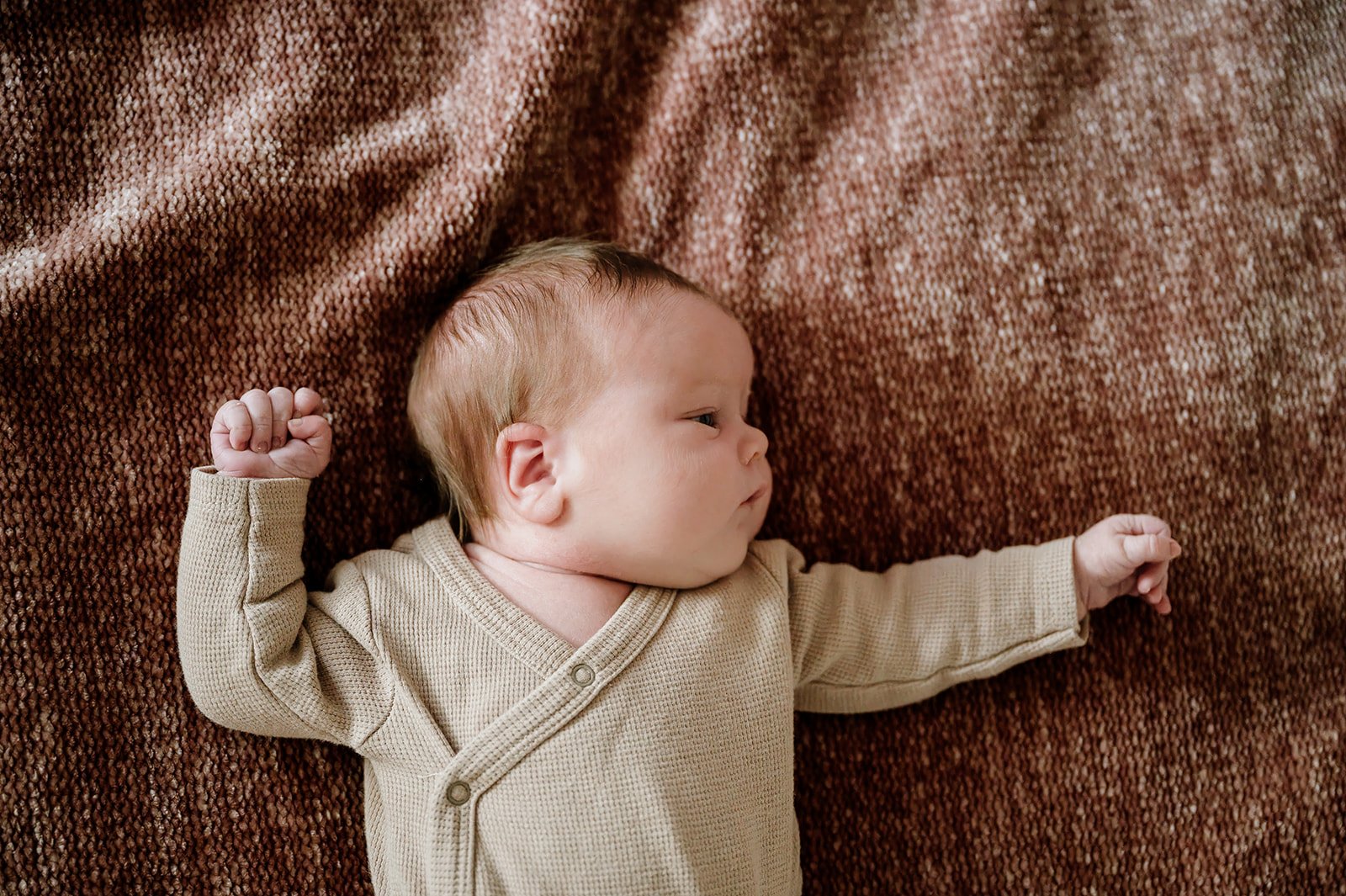 A baby lying on a textured brown blanket, sleeping peacefully with arms stretched out.