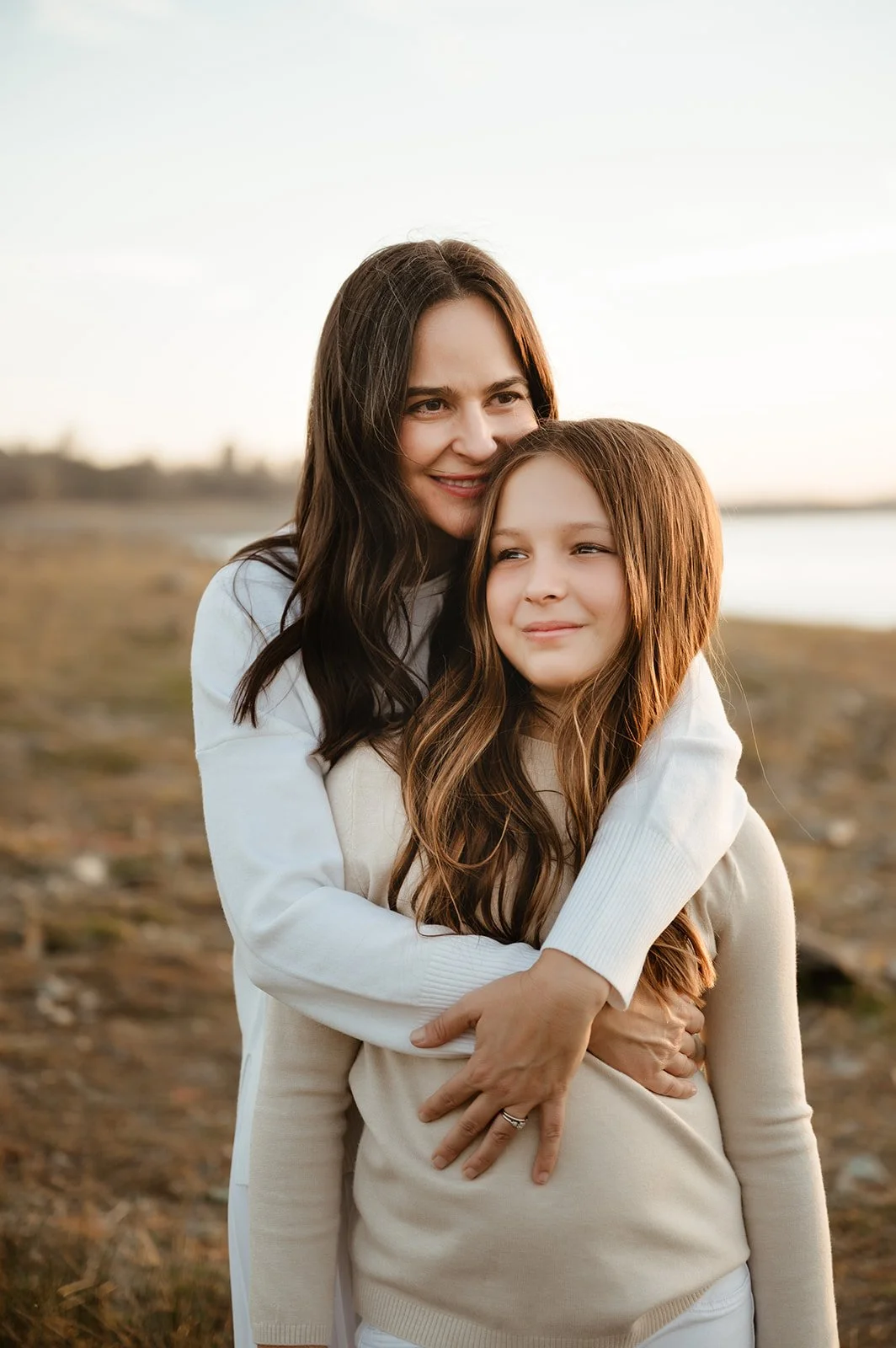 A woman hugging a girl outdoors near a body of water during sunset.