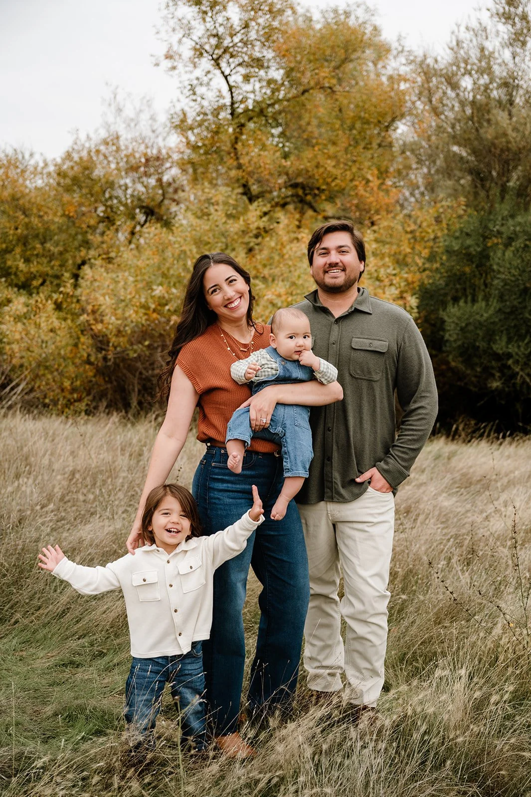 A family of four standing outdoors in a grassy field with autumn trees in the background. The mother holds a baby, while the father stands beside her, and a young girl stands in front.