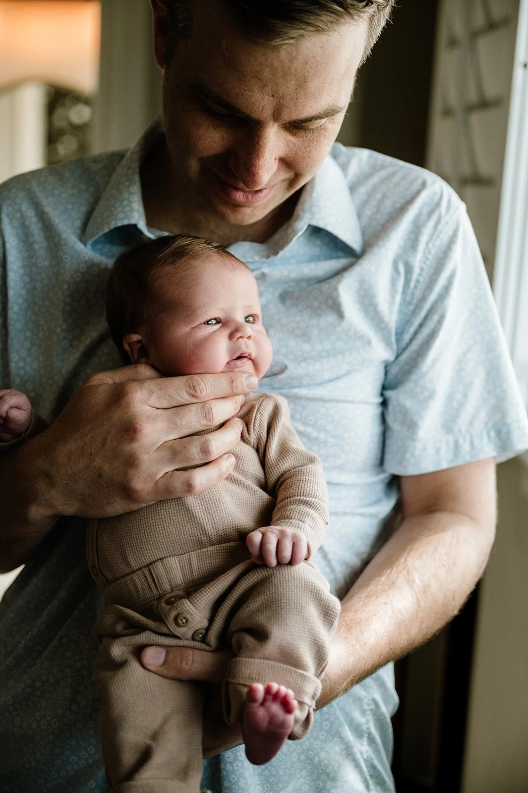A man holding a newborn baby near a window, looking down at the baby with a gentle smile.