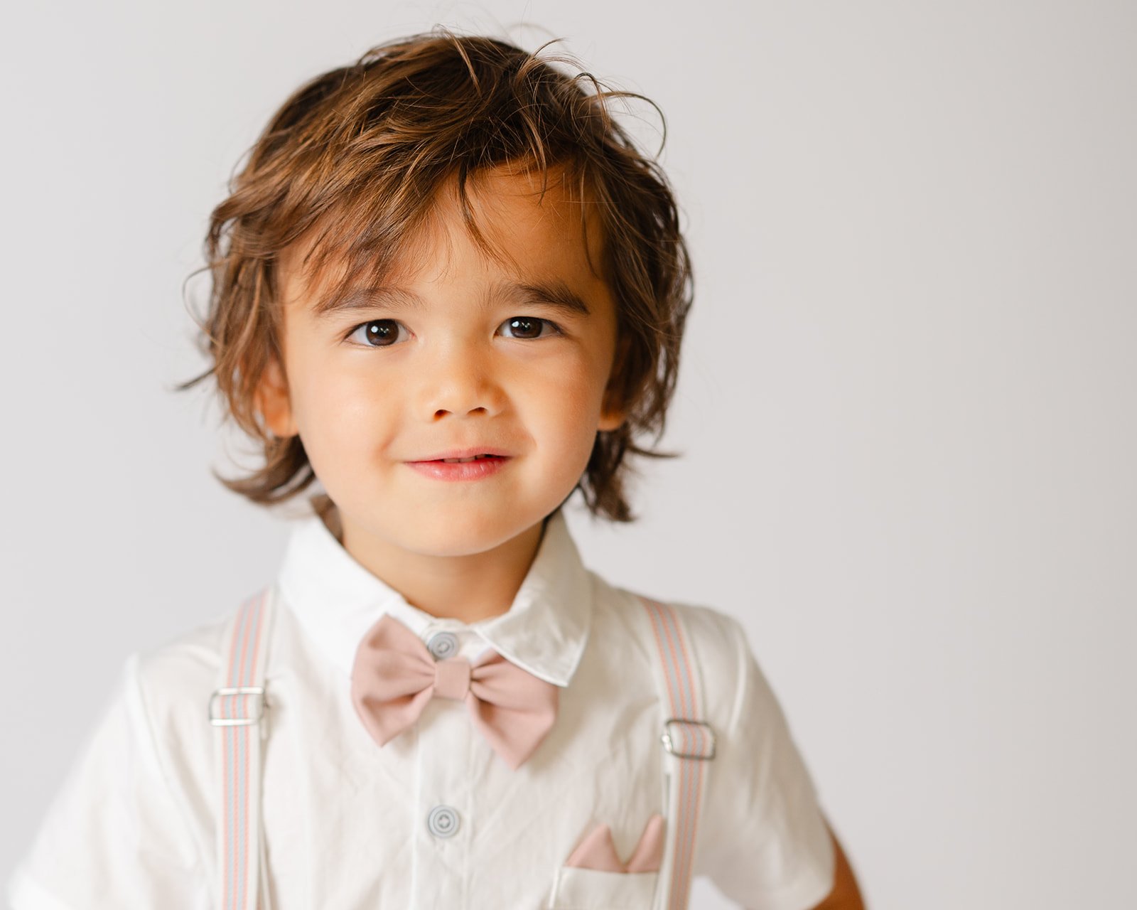 A young boy with brown, curly hair, wearing a white shirt with a pink bow tie, suspenders, and a pocket square, smiling at the camera against a plain light gray background.