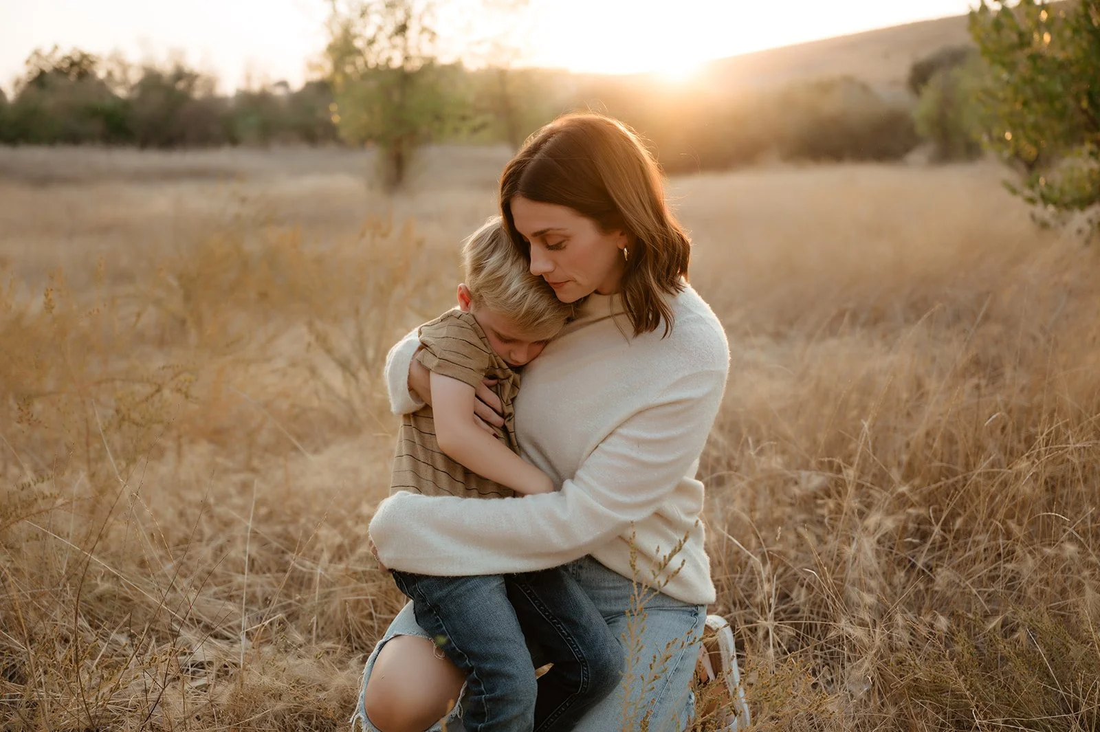 A woman cuddling a young boy in a field of tall dry grass with trees in the background during sunset.