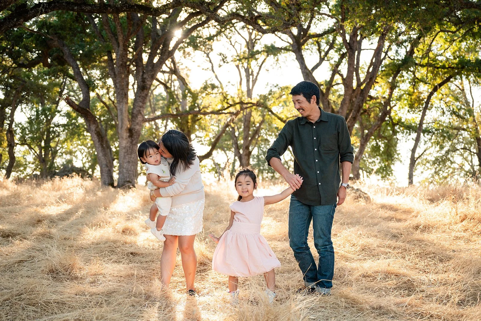 Happy family of four enjoying a walk in a sunlit, grassy park with large trees, holding hands and smiling.