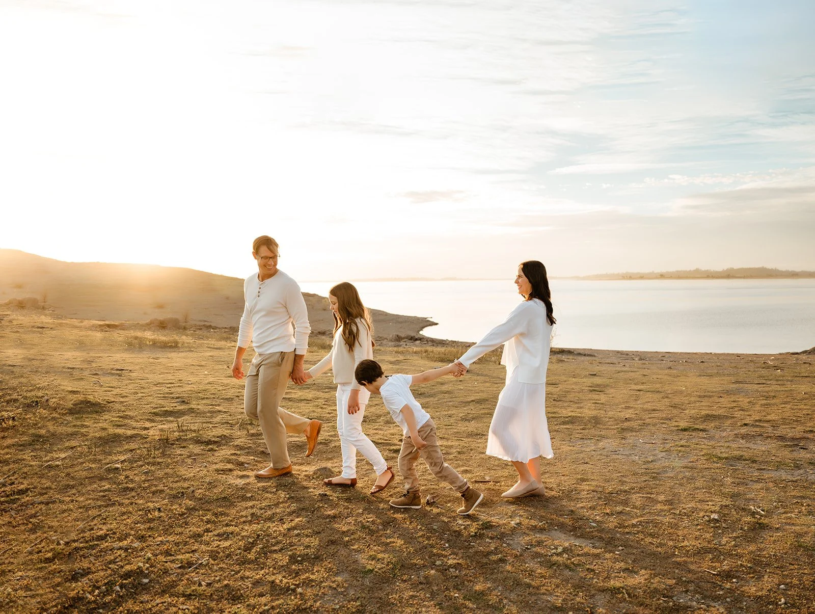 Family of five walking hand in hand along a lakeshore at sunset, with a man, woman, two children, and a young girl, all dressed in neutral tones and smiling.