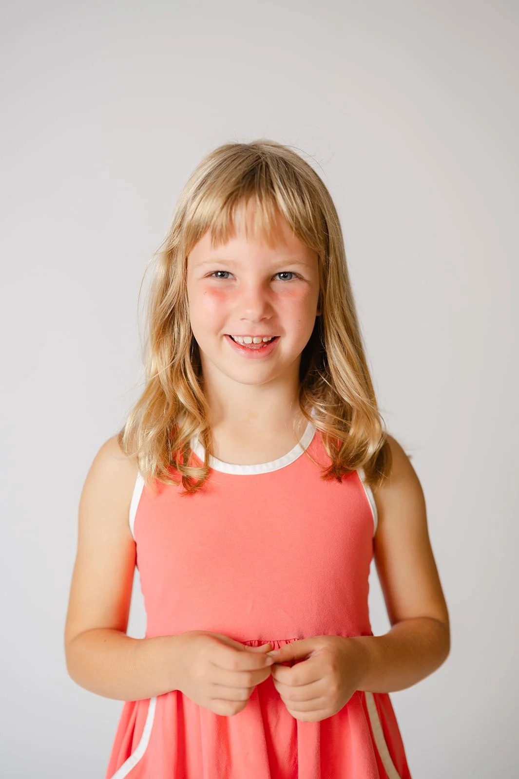 A young girl with blonde hair wearing a coral dress with white trim, smiling and standing against a plain white background.