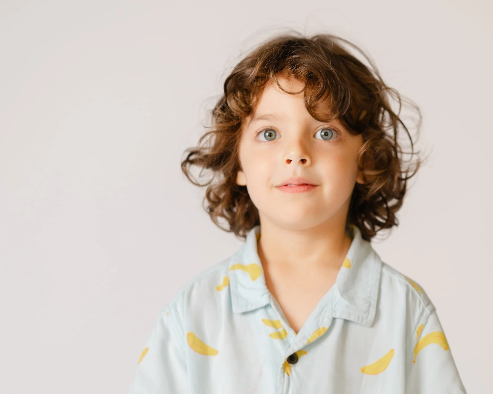 A young boy with curly brown hair and blue eyes wearing a light-colored shirt with yellow banana prints, looking directly at the camera with a slight smile.