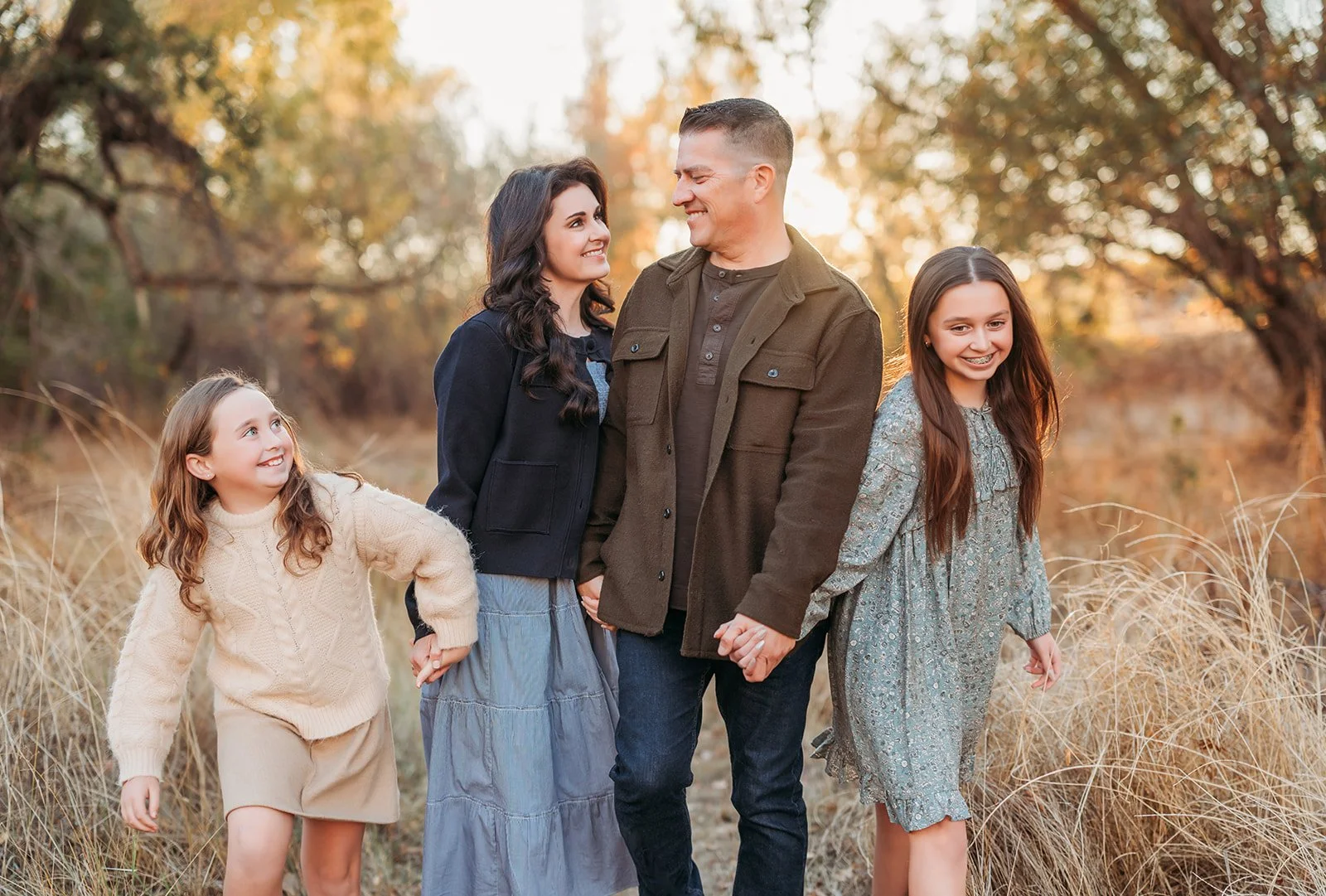 Family of four holding hands and walking outdoors in fall foliage, smiling at each other.
