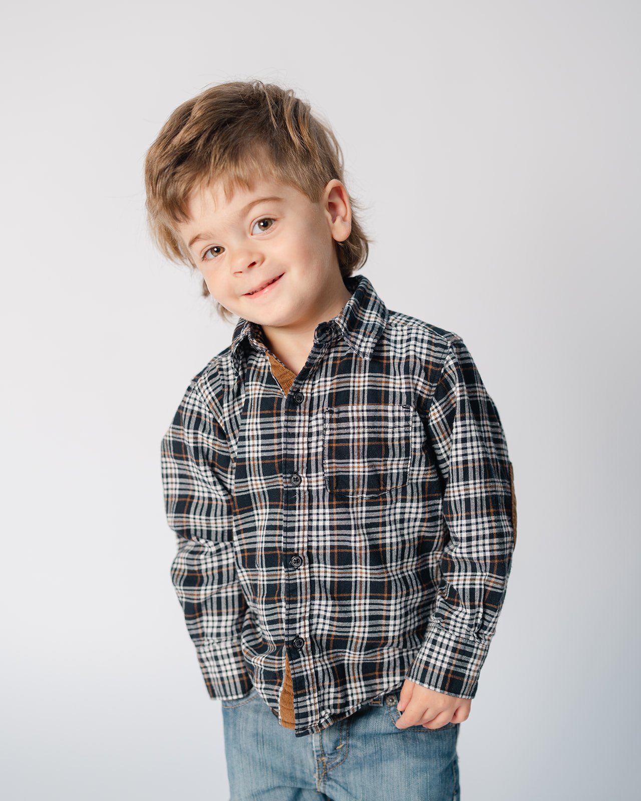 Young boy with light brown hair and light skin wearing a plaid button-up shirt and blue jeans standing against a plain light gray background, smiling with a tilted head.