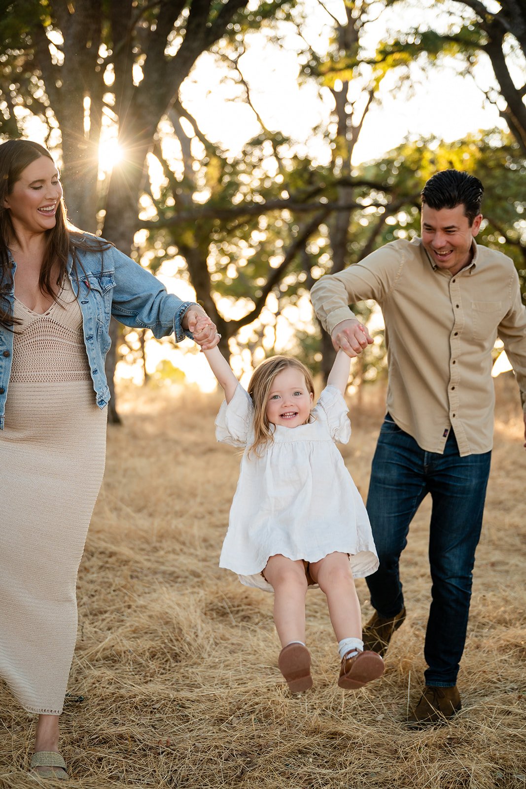 A happy family of three, a mother, father, and young daughter, holding hands and swinging in a field of dry grass during sunset. Trees and sunlight are in the background.