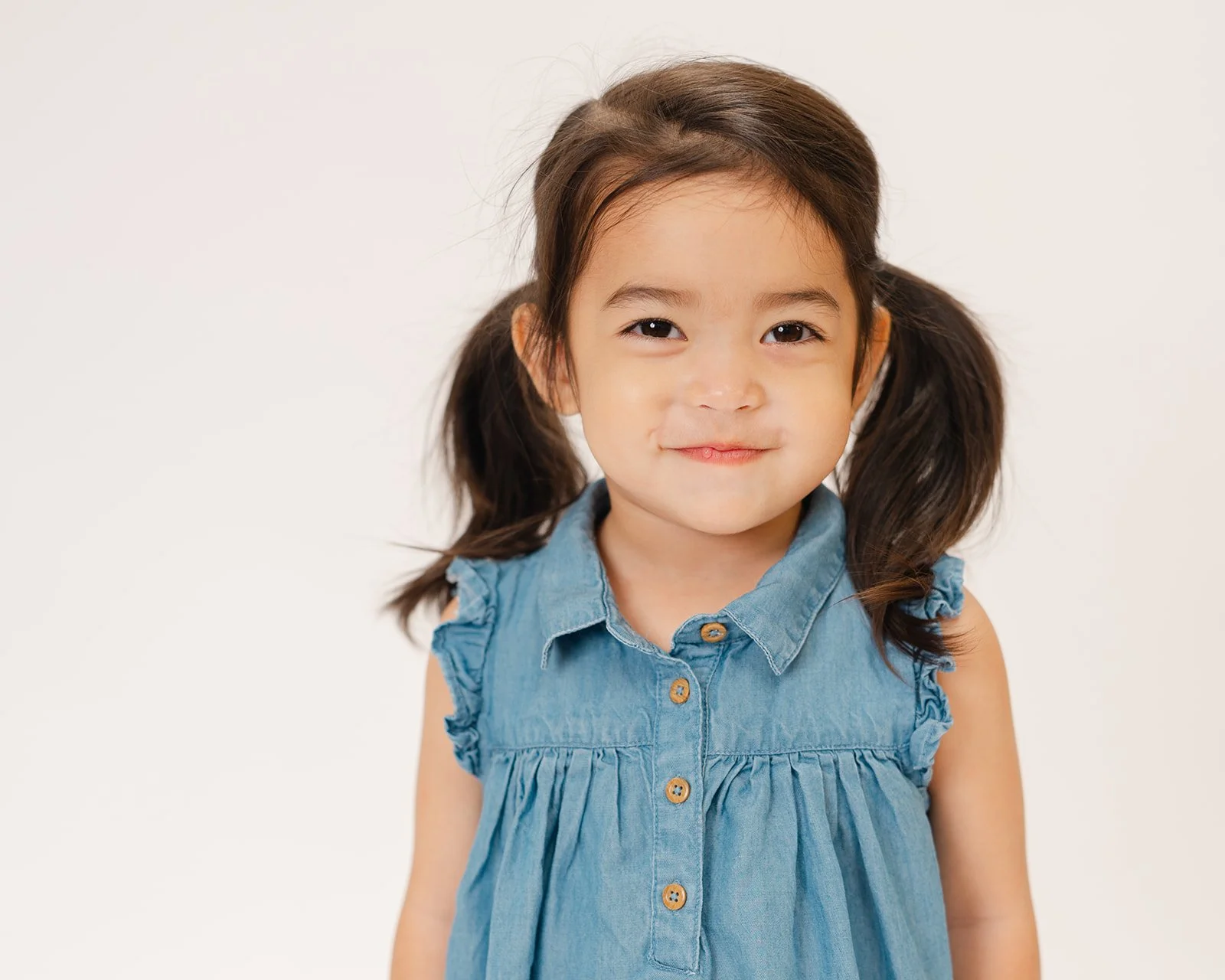 A young girl with dark brown hair in pigtails wearing a sleeveless denim dress, smiling at the camera.