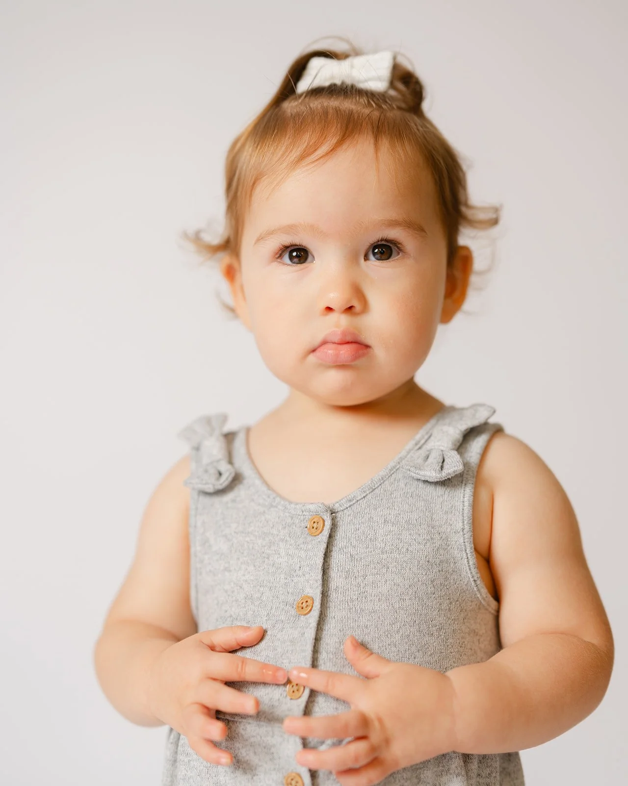 A young girl with light skin, brown eyes, and reddish hair tied with a white bow, wearing a gray sleeveless dress with button details and ruffled shoulders, standing against a plain white background.