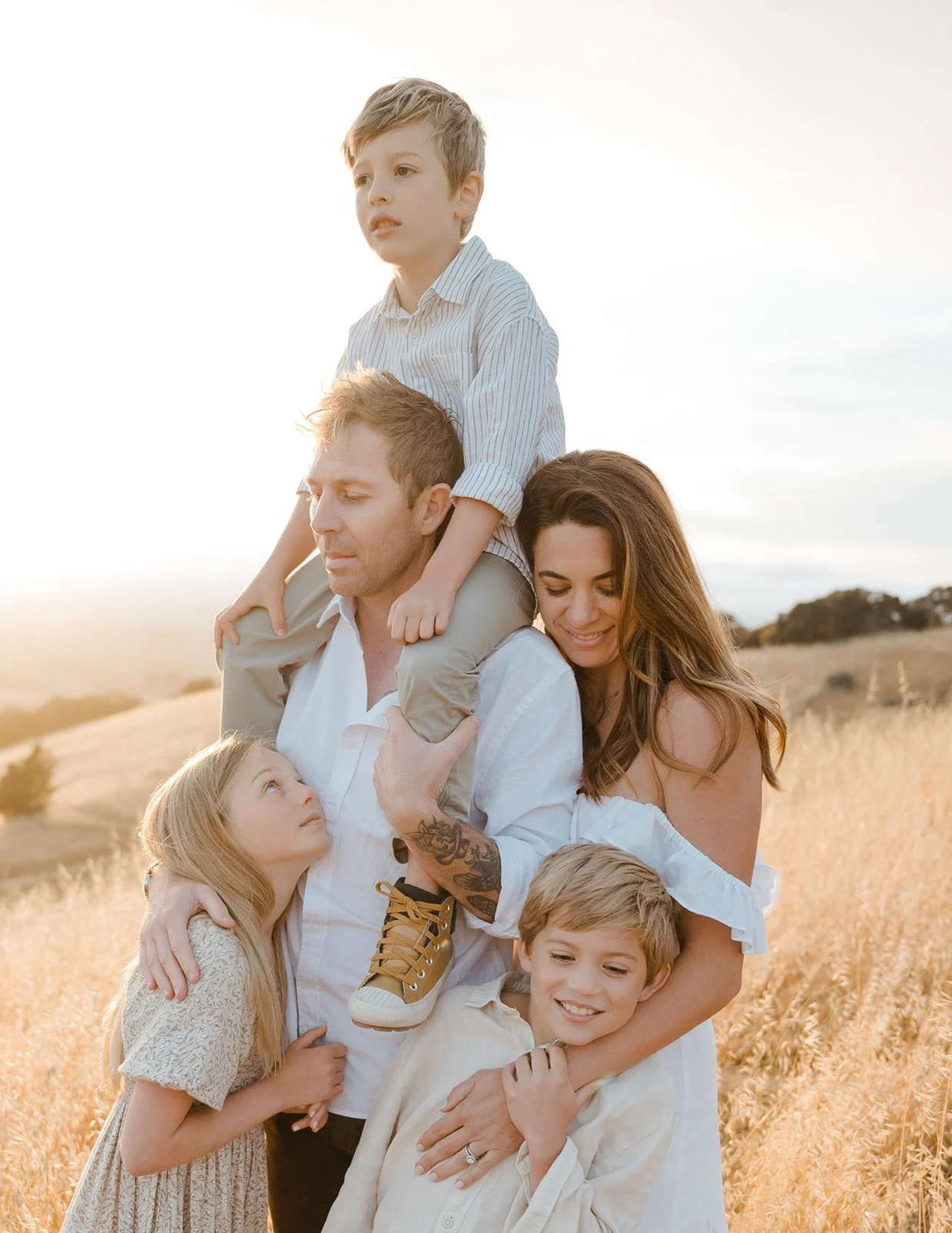 A family of six outdoors in a field during sunset, with the father carrying a young boy on his shoulders, the mother hugging a young girl, and the older boy smiling at the camera.