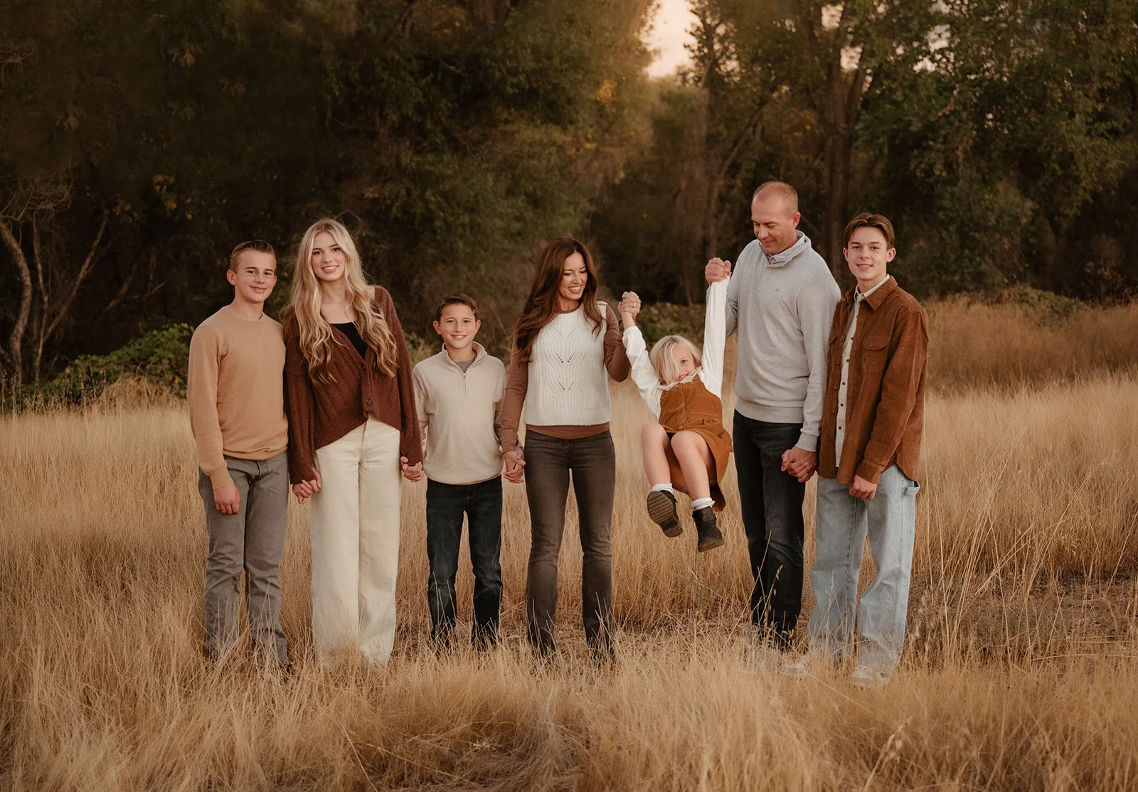 Family of eight holding hands in a grassy field during fall, with trees in the background. One child is being lifted off the ground by two adults.