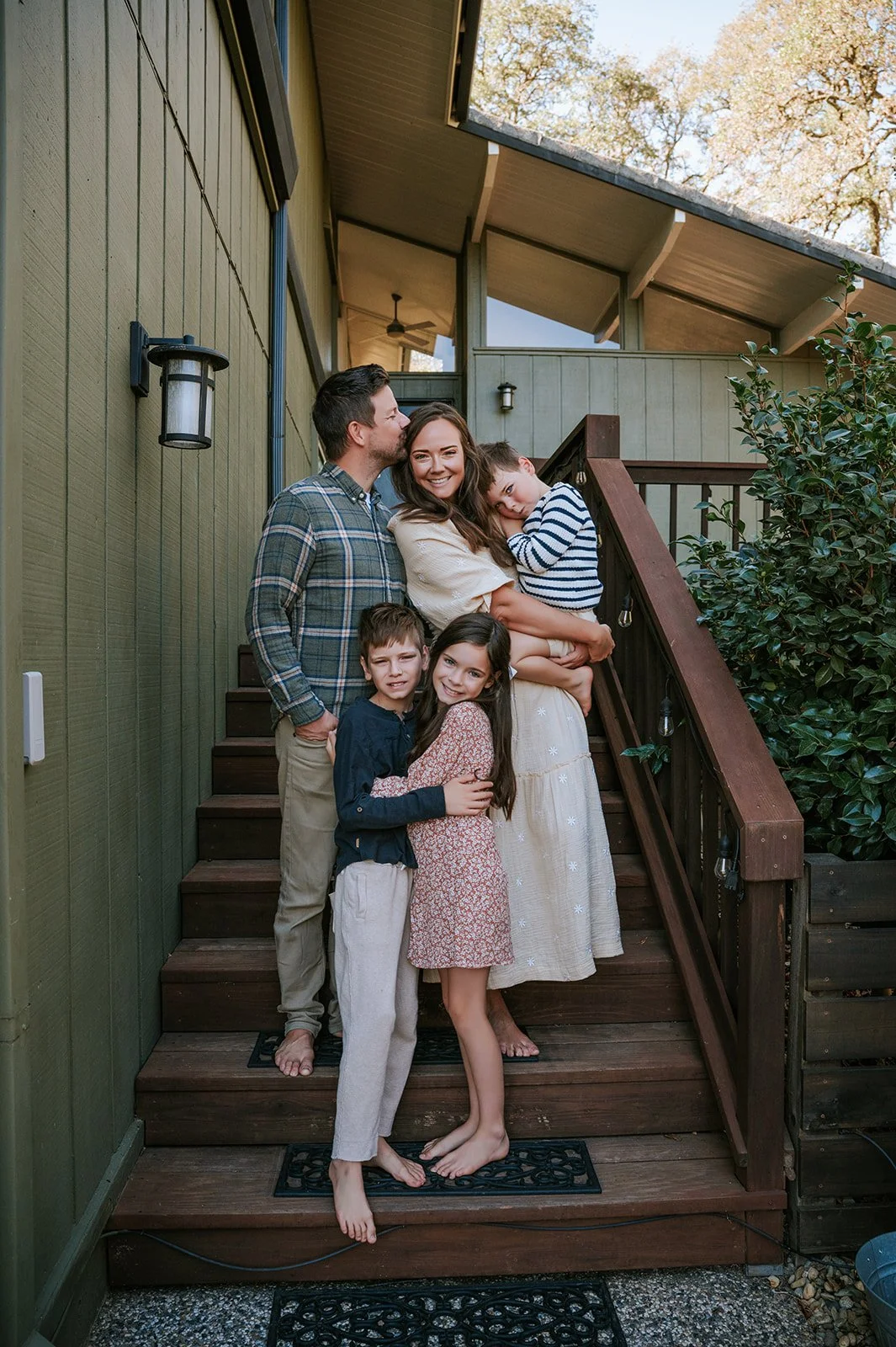A family of five stands on a wooden porch steps outside a house. The family includes a man, woman, and three children. The woman is holding a young boy in striped shirt, and the other two children are hugging the woman. The family is smiling, and the