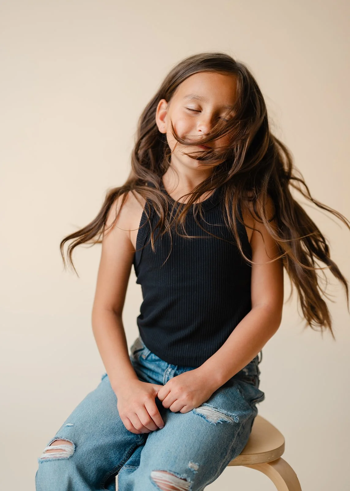 A young girl with long brown hair sitting on a wooden stool against a plain beige background, wearing a black sleeveless top and ripped blue jeans, with her eyes closed and hair blowing across her face.