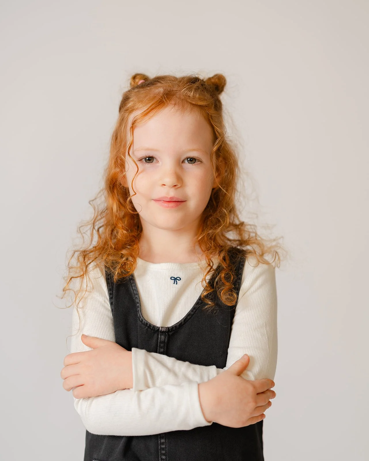 A young girl with curly red hair styled in pigtails, wearing a cream long-sleeve shirt and a black dress, standing with her arms crossed against a light background.