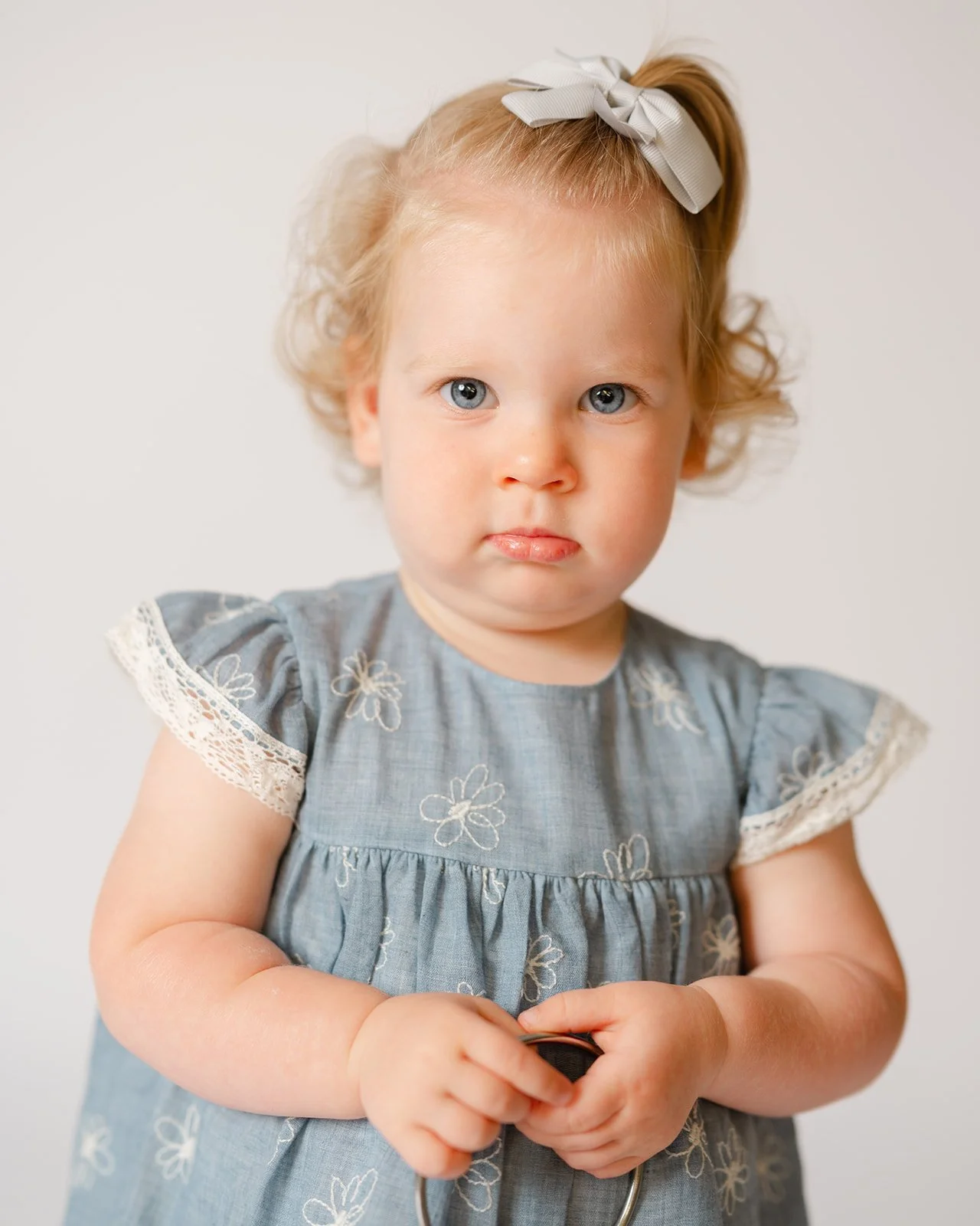 A young girl with blue eyes and blonde curly hair tied with a gray bow, wearing a gray dress with floral embroidery and lace trim, standing against a white background.