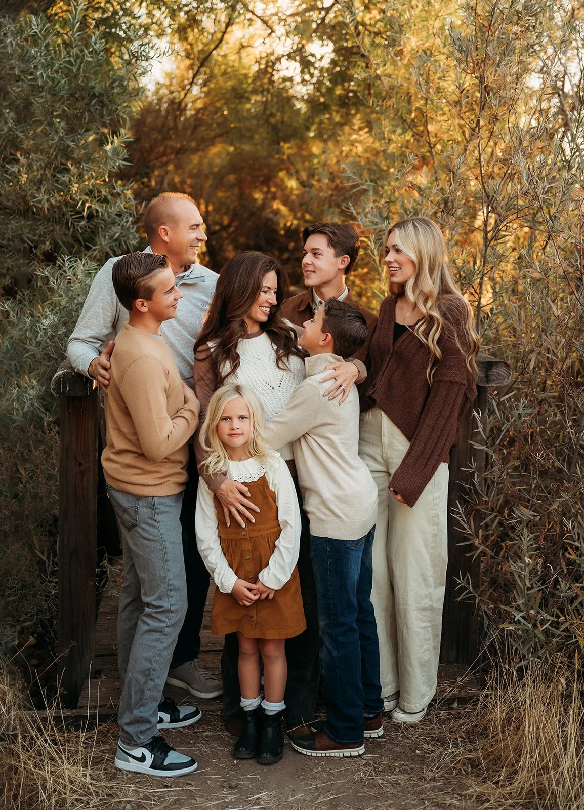 A multigenerational family of nine enjoying an outdoor moment during fall, surrounded by trees with autumn foliage, standing on a wooden bridge.