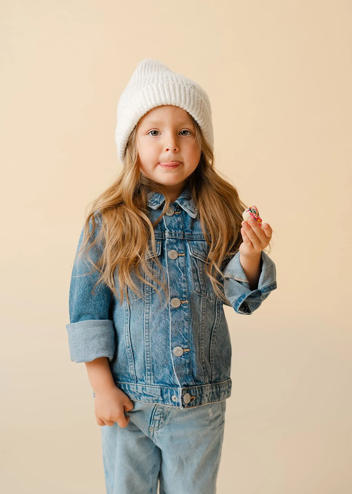 A young girl with long blonde hair wearing a white knit beanie, a denim jacket, and light jeans, holding a decorated cupcake with pink frosting.
