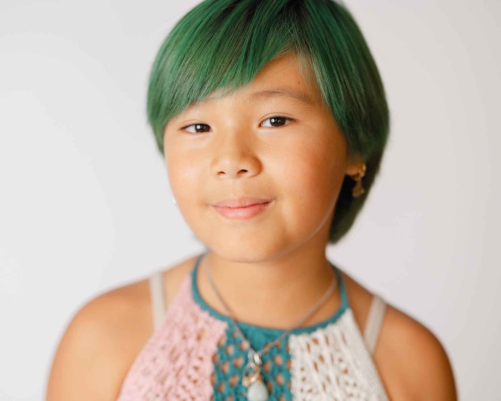 A young girl with green hair and a gentle smile, wearing earrings, layered necklaces, and a sleeveless crochet top, against a plain white background.