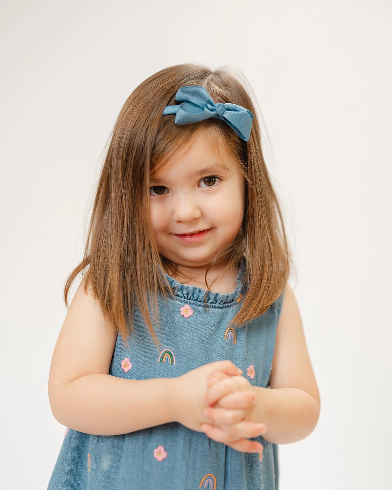 Young girl with light brown hair wearing a blue dress with rainbow and flower embroidery, a blue headband with a bow, smiling and clasping her hands together against a plain white background.