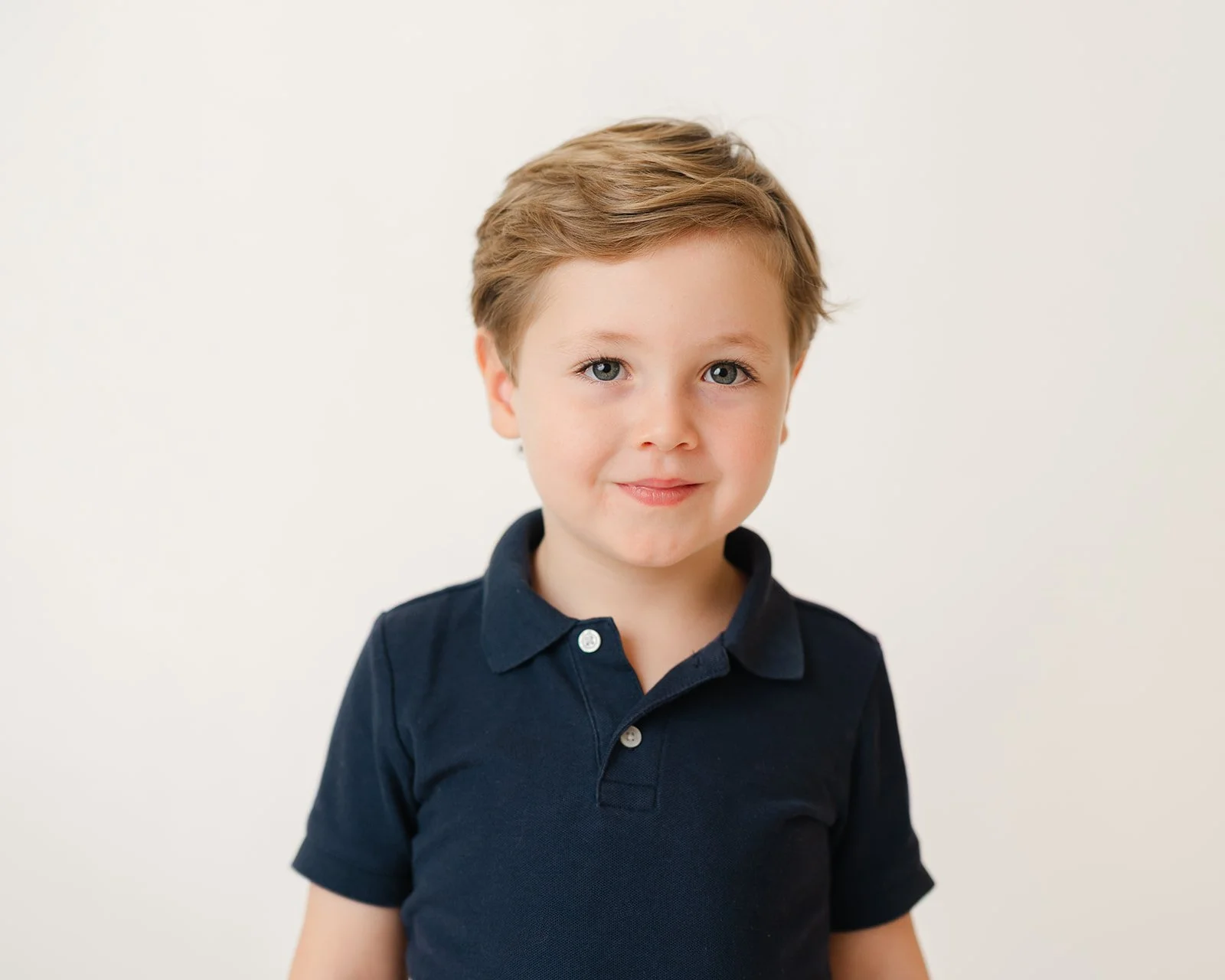 A young boy with light brown hair, blue eyes, and a slight smile, wearing a dark blue collared shirt, standing against a plain white background.