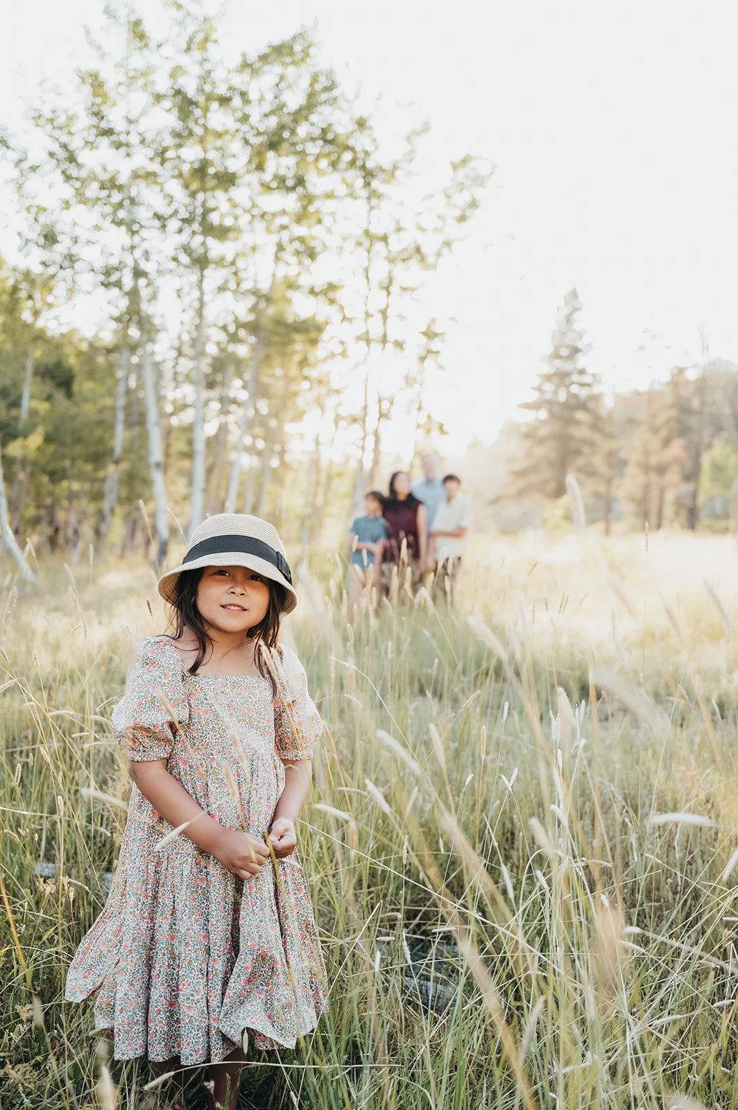 A young girl in a floral dress and a sun hat standing in a grassy field with a family in the background.
