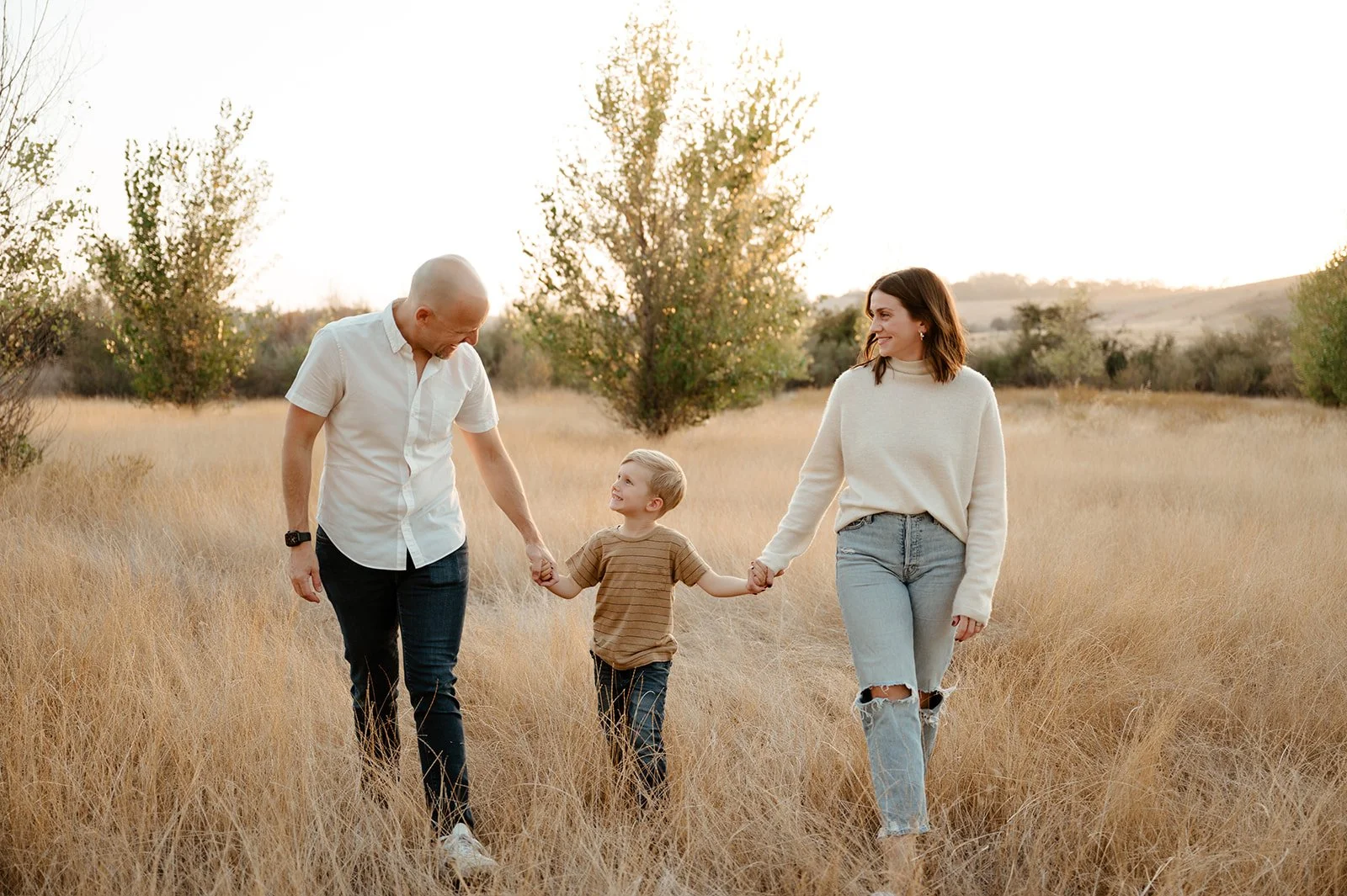 Family of three walking through a grassy field holding hands, with trees and hills in the background during sunset.