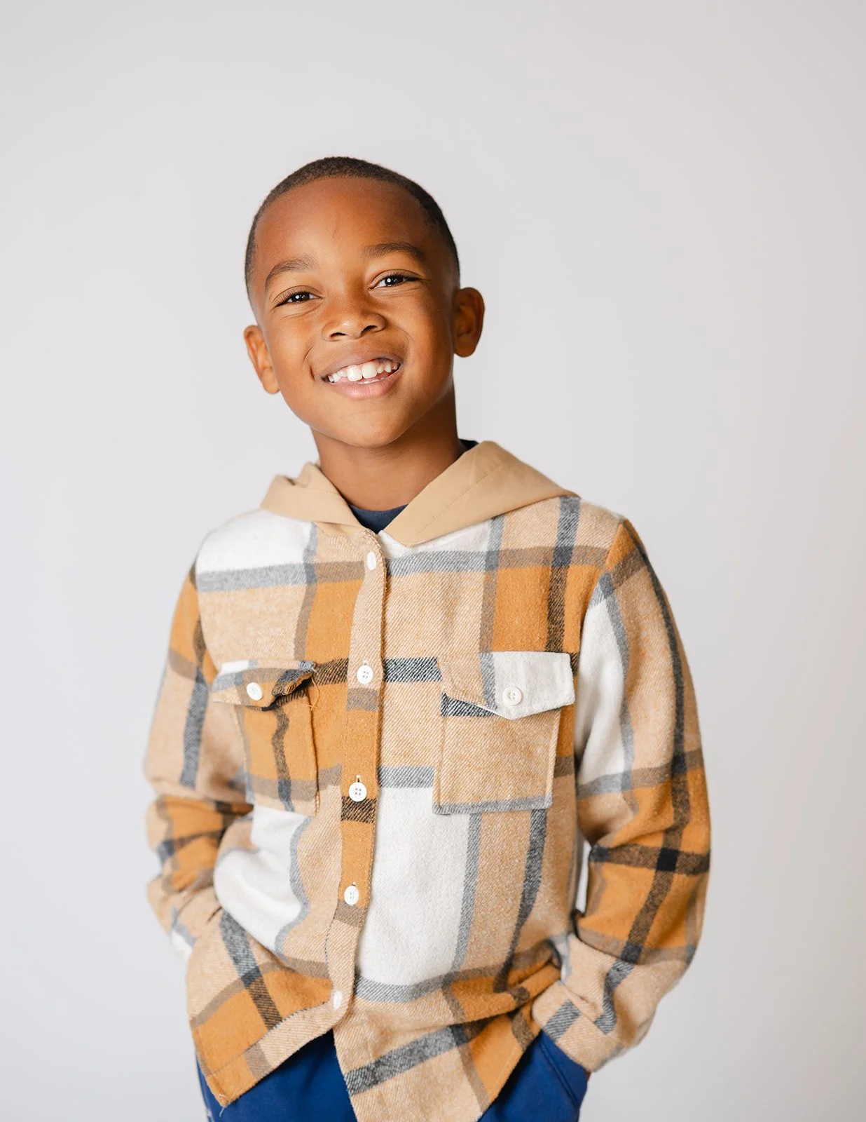 A smiling young boy with dark skin, wearing a tan and white plaid button-up shirt over a beige hoodie, set against a plain white background.