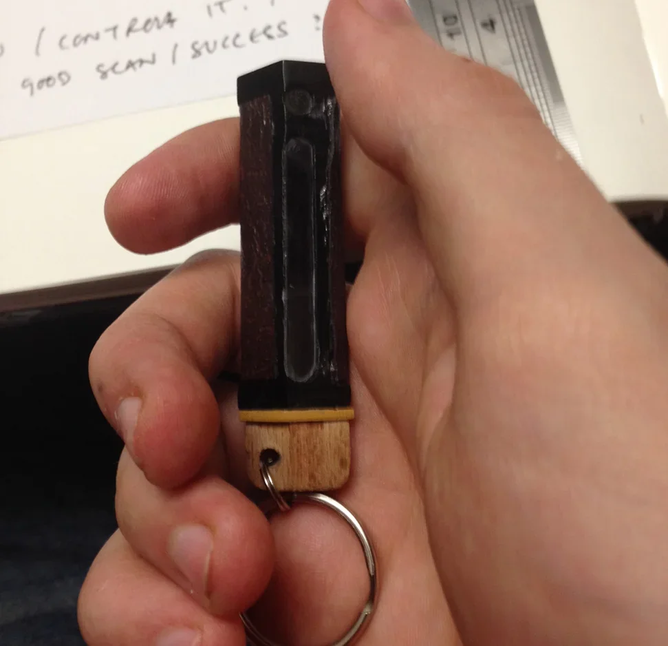 Person holding a small wooden and black plastic device with a metal keyring attached at the bottom.