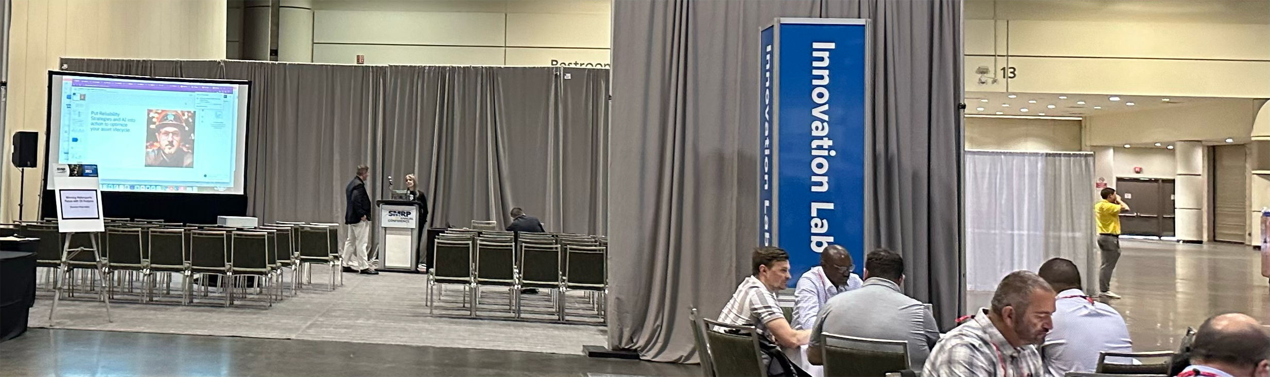 A conference room with a stage and a large screen displaying a presentation. There are several chairs arranged in rows, some occupied by people, and others empty. Two people are standing at a podium, and a person is seated at a table near the stage. The backdrop is made of gray curtains, and a tall blue sign reads 'Innovation Lab'.