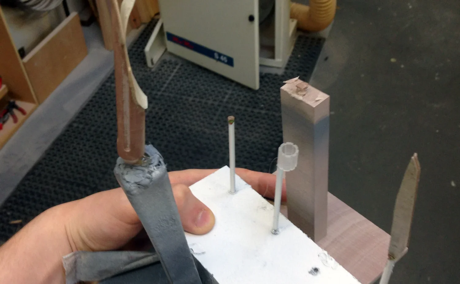 Close-up of a hand holding a small handmade model of a UFO with metal and foam parts, with various thin rods protruding from the base, in a workshop or craft space.