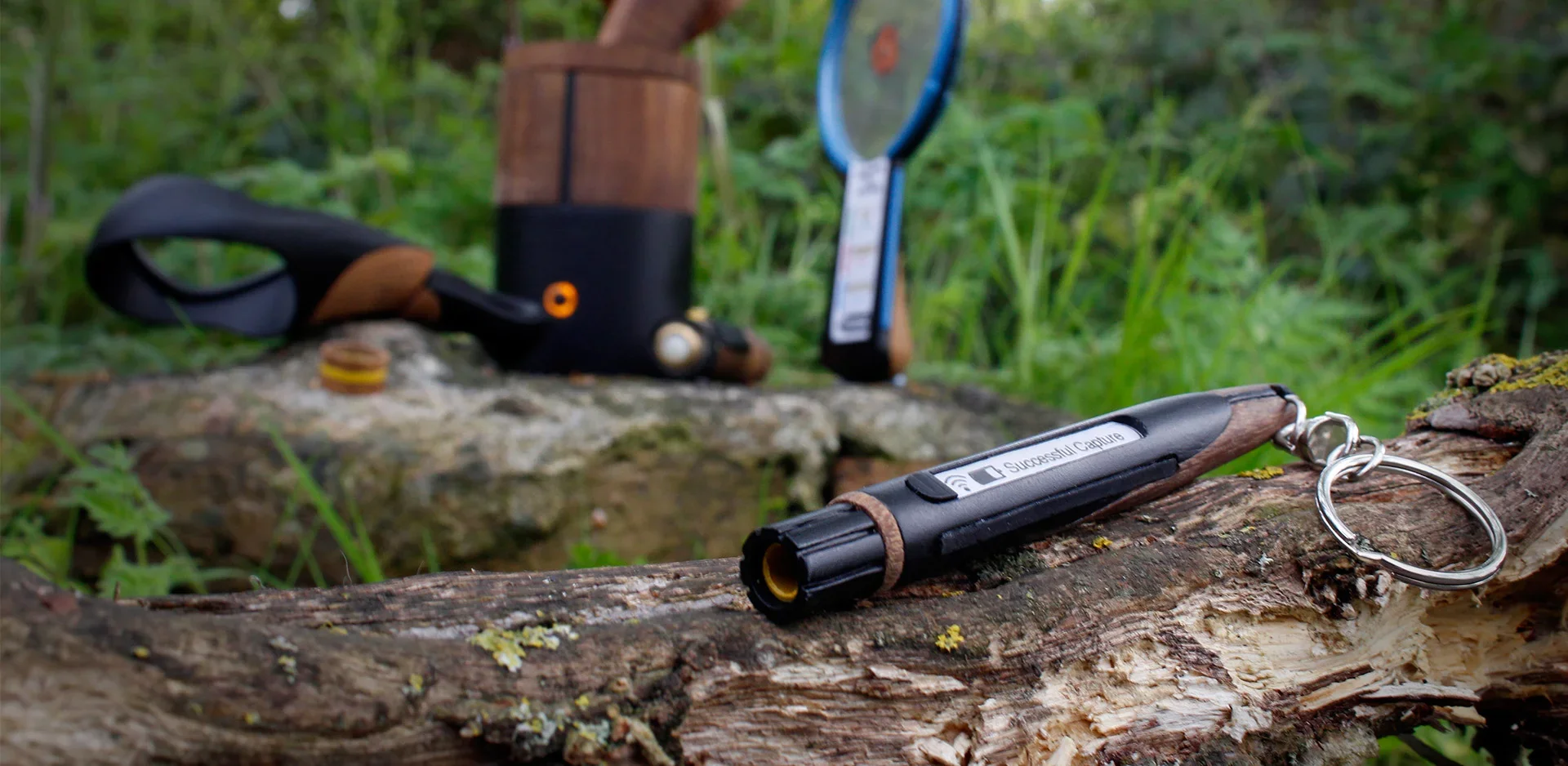 Close-up of a keychain LED flashlight with a metal loop on a fallen tree in a forest, with additional blurred outdoor gear in the background.