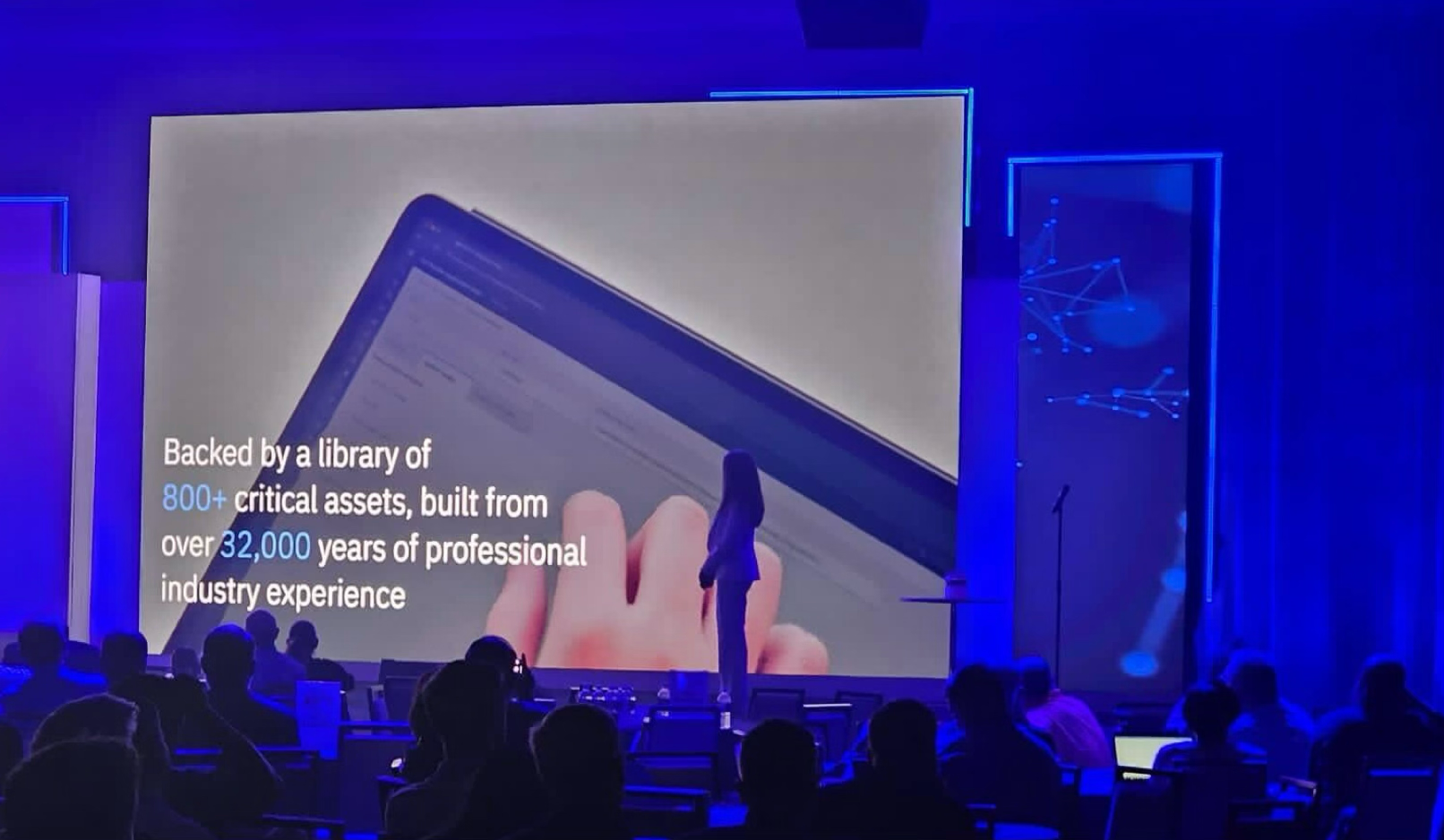 At a conference, a speaker stands in front of a large screen displaying text that reads: "Backed by a library of 800+ critical assets, built from over 32,000 years of professional industry experience." Audience members are seated, watching the presentation.