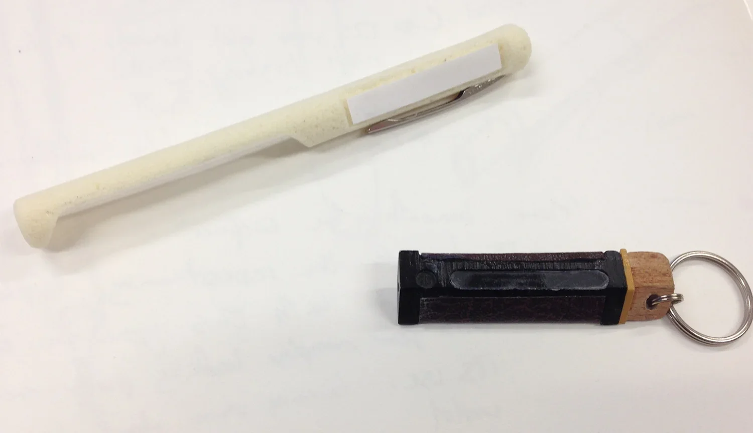 A white pen-shaped tool and a wooden and black keychain with a small rectangular shape are on a white surface.